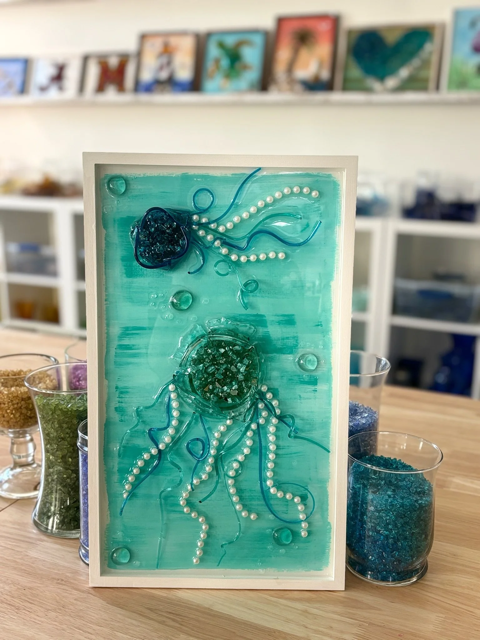 A framed resin artwork of ocean-themed art featuring a jellyfish design using pearls and blue strings, and other glass.