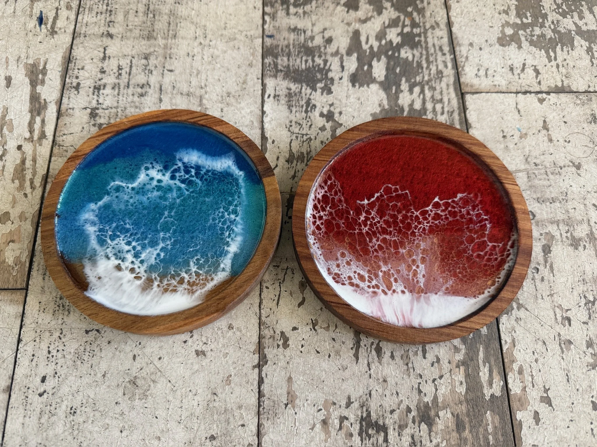 Two round wooden coasters containing colorful resin art, one blue with white swirling patterns and one red with white ocean wave patterns.