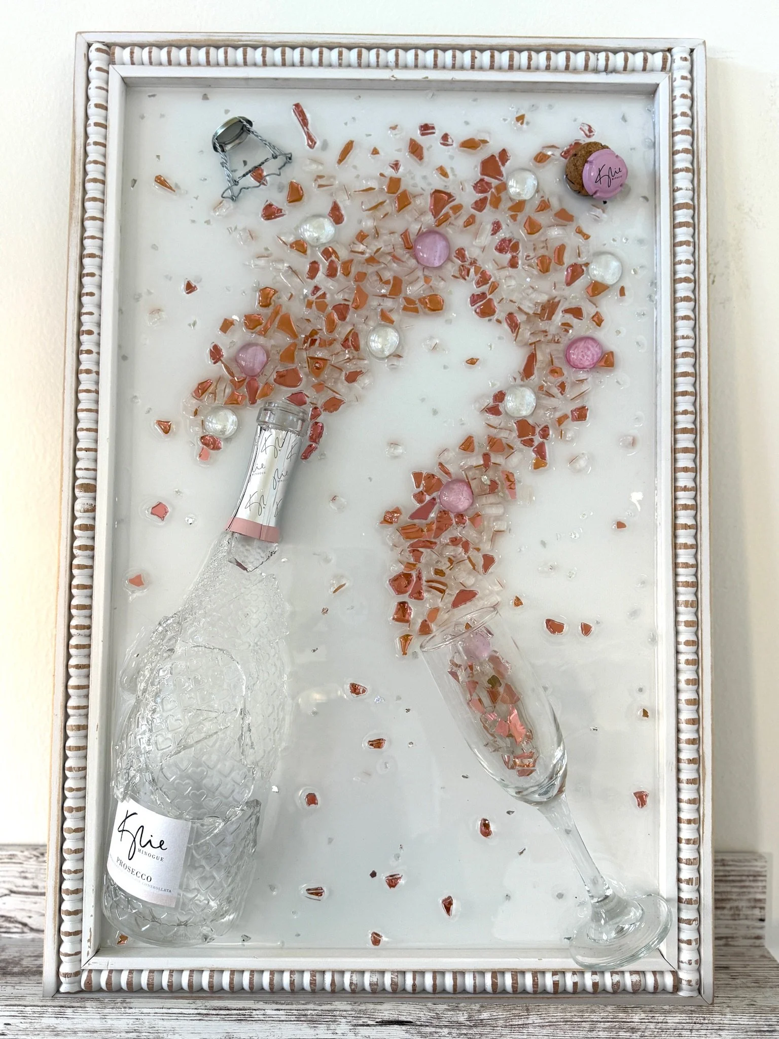 Resin Glass Art of cut and crushed glass. Broken rose bottle with pink, clear, and brown glass pieces, two empty glass flutes, and scattered pink and pearlescent beads. Man Cave Art - Home Bar - She Shed.