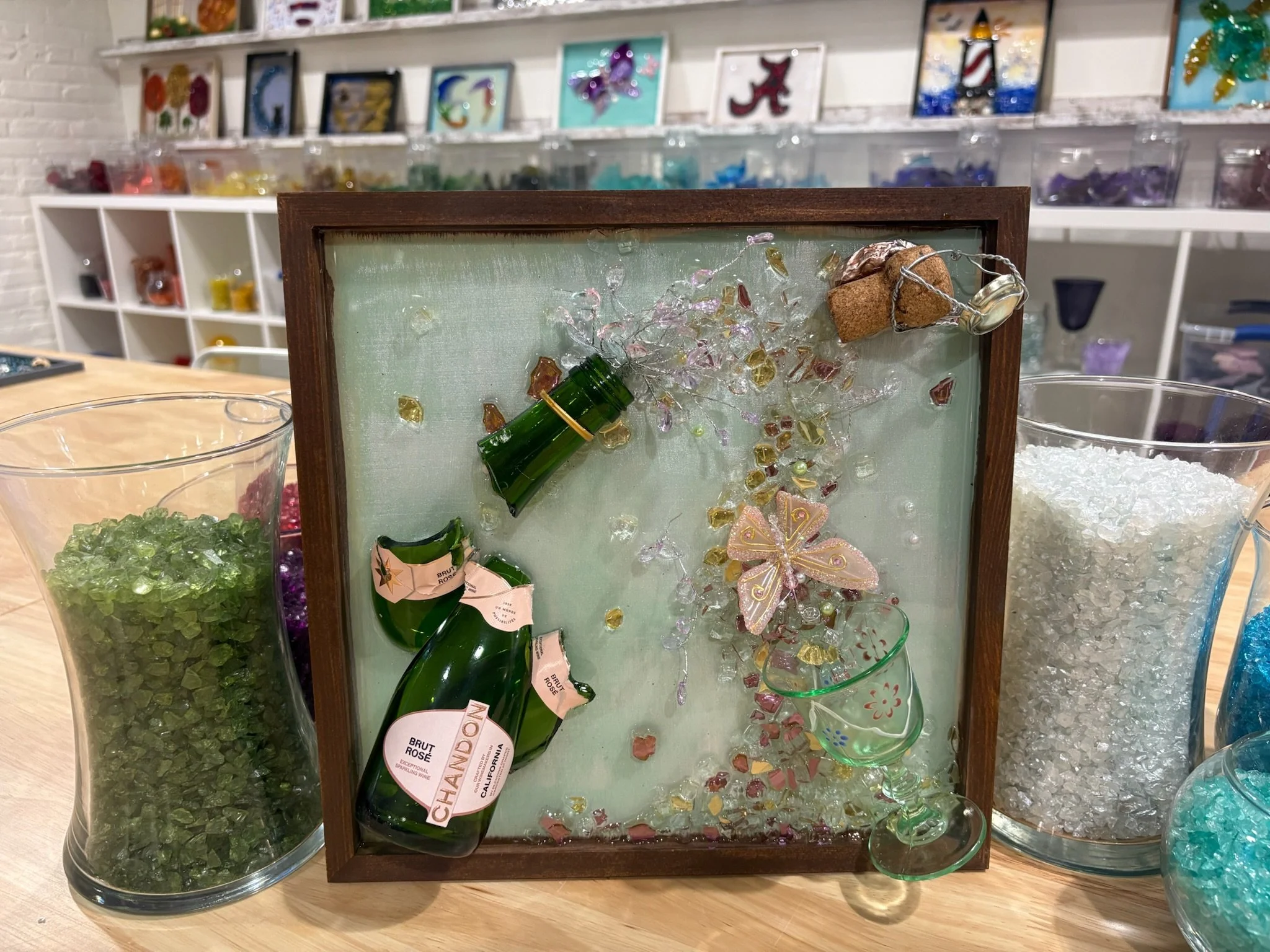 Resin art using cut and crushed glass of a small broken champagne glass and ceramic pieces and a miniature green champagne bottle in a painted frame.