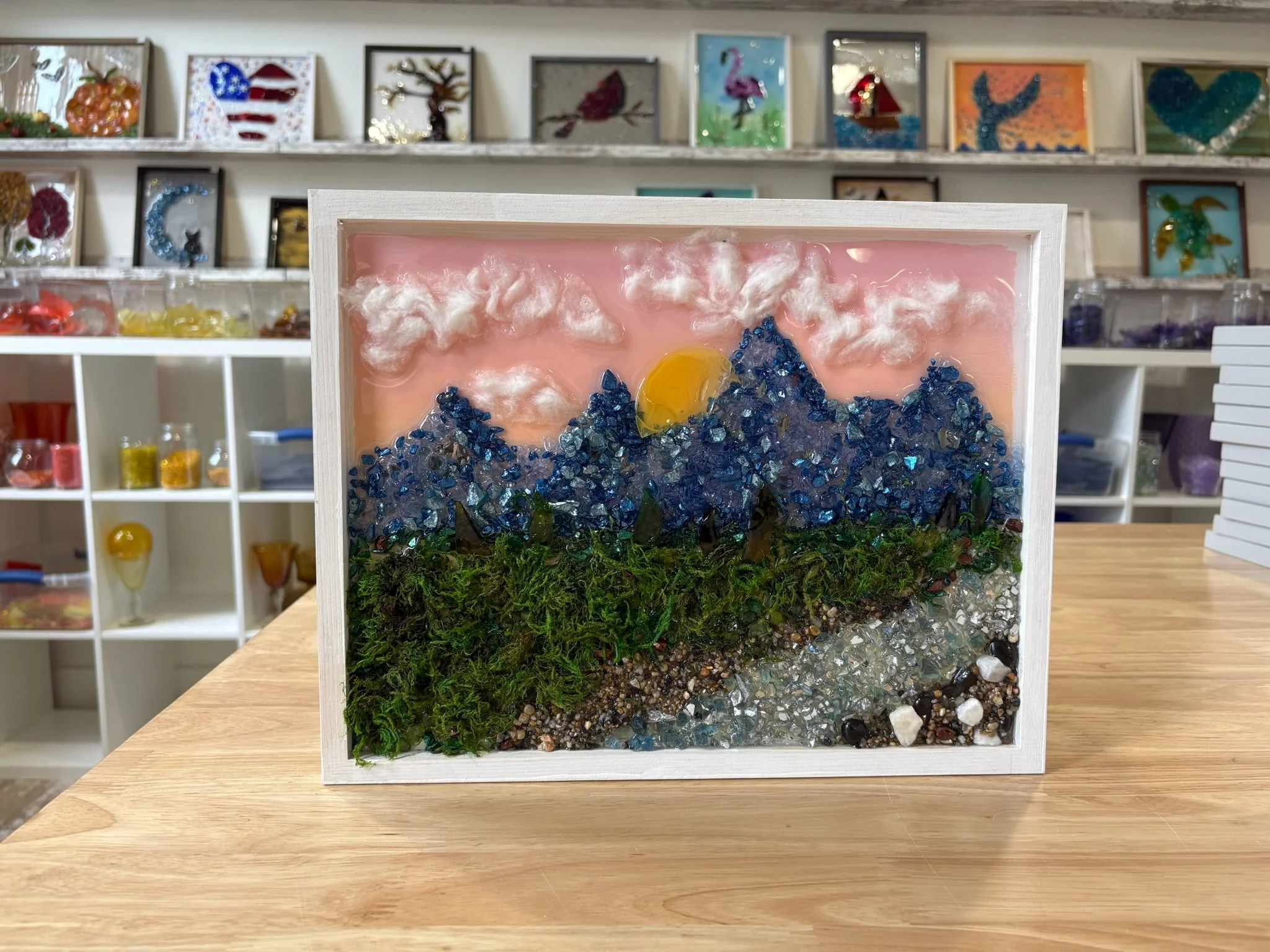 A framed resin artwork using cut and crushed glass featuring a sunset scene with mountains, clouds, and a pink sky, displayed on a wooden table in front of a wall with various colorful art pieces in glass frames.
