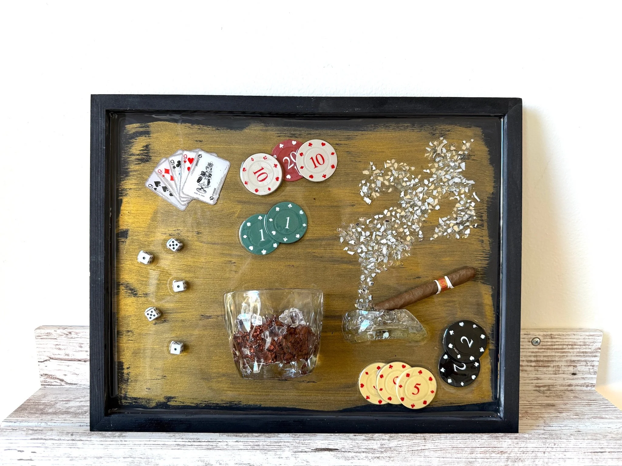 A rectangular wooden tray with a black border containing playing cards, poker chips, dice, a glass of ice, dried berries, and a cigar.