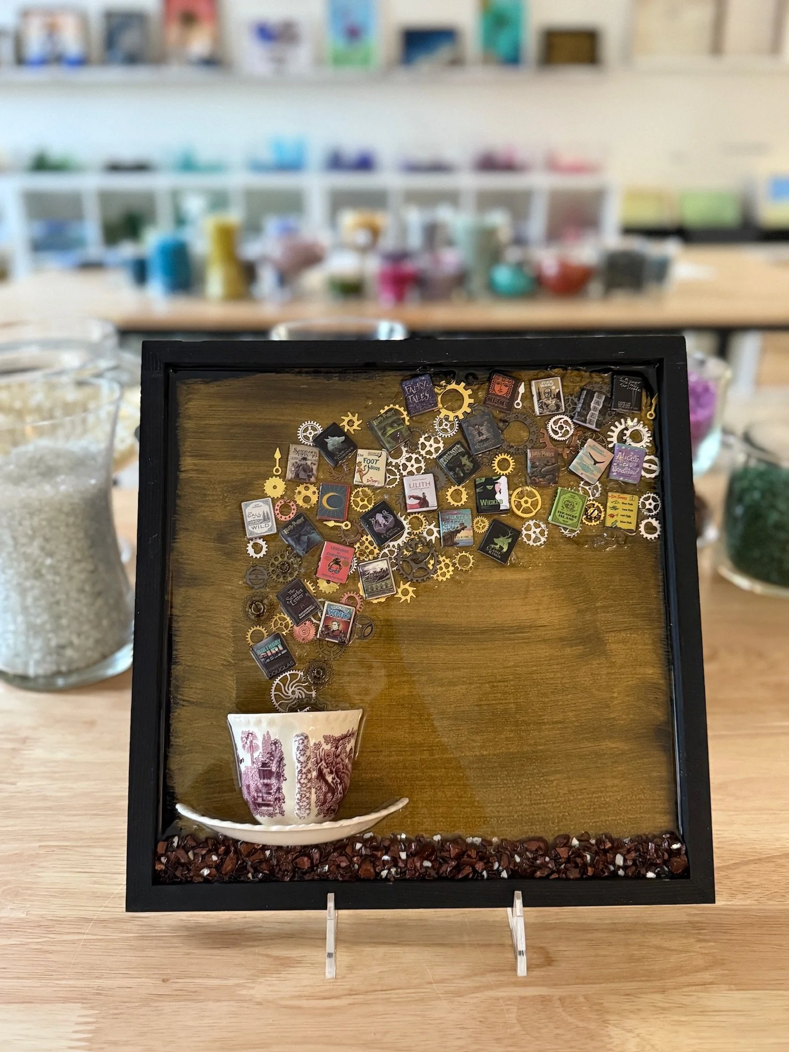 Resin Glass Art of a tea cup with miniature books flowing in/out of it using cut and crushed glass in a painted frame.