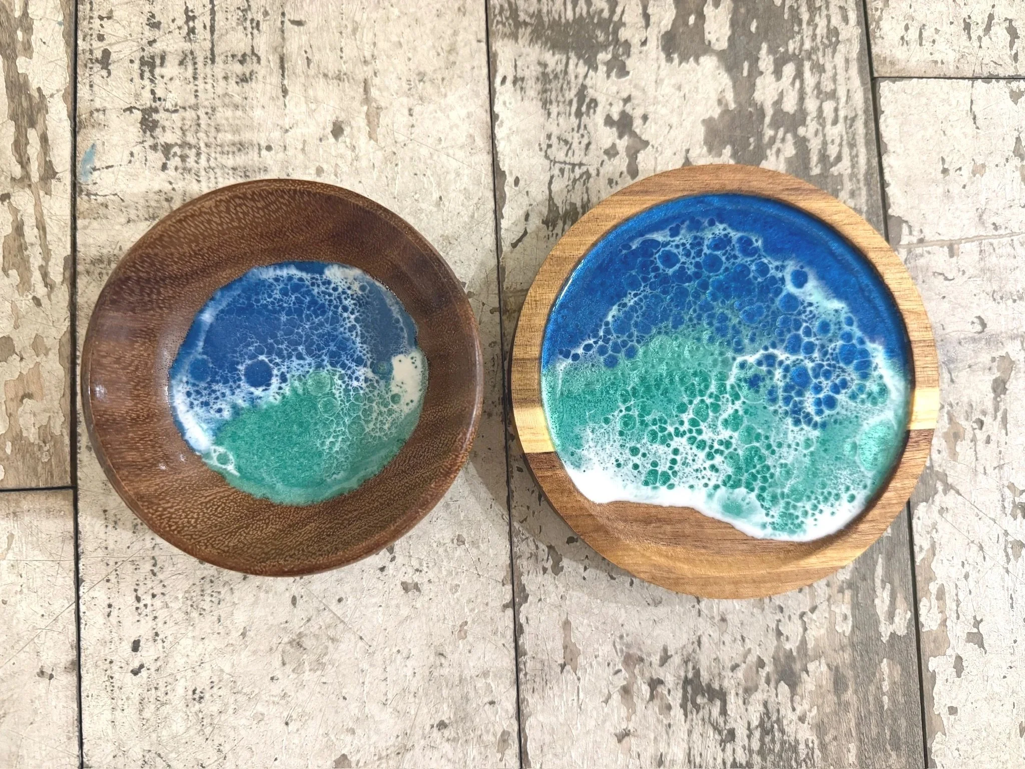 Two round wooden bowls with ocean-like resin art, featuring blue, white, and green swirls, placed on a distressed wooden surface.