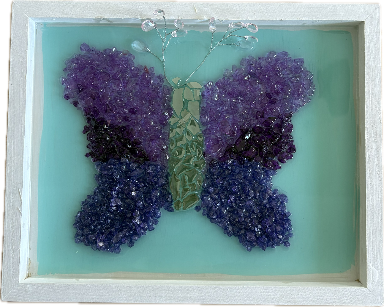 Framed resin art of a butterfly made from purple glass stones and green glass pieces with wire antennae, set against a light turquoise background.