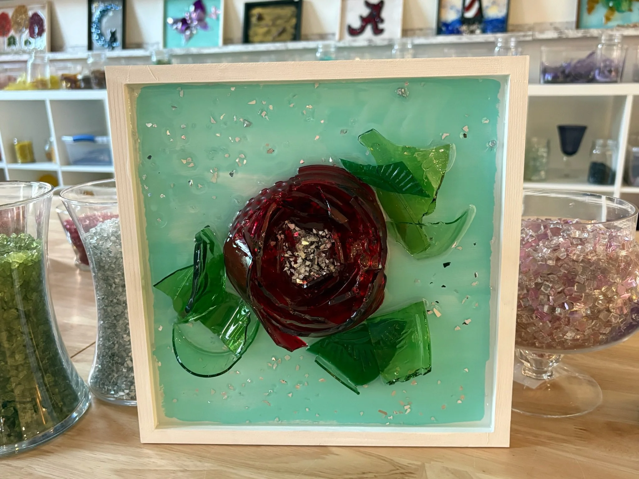 Resin Glass artwork using cut and crushed glass making a red rose with green leaves on a light blue background, framed in wood.