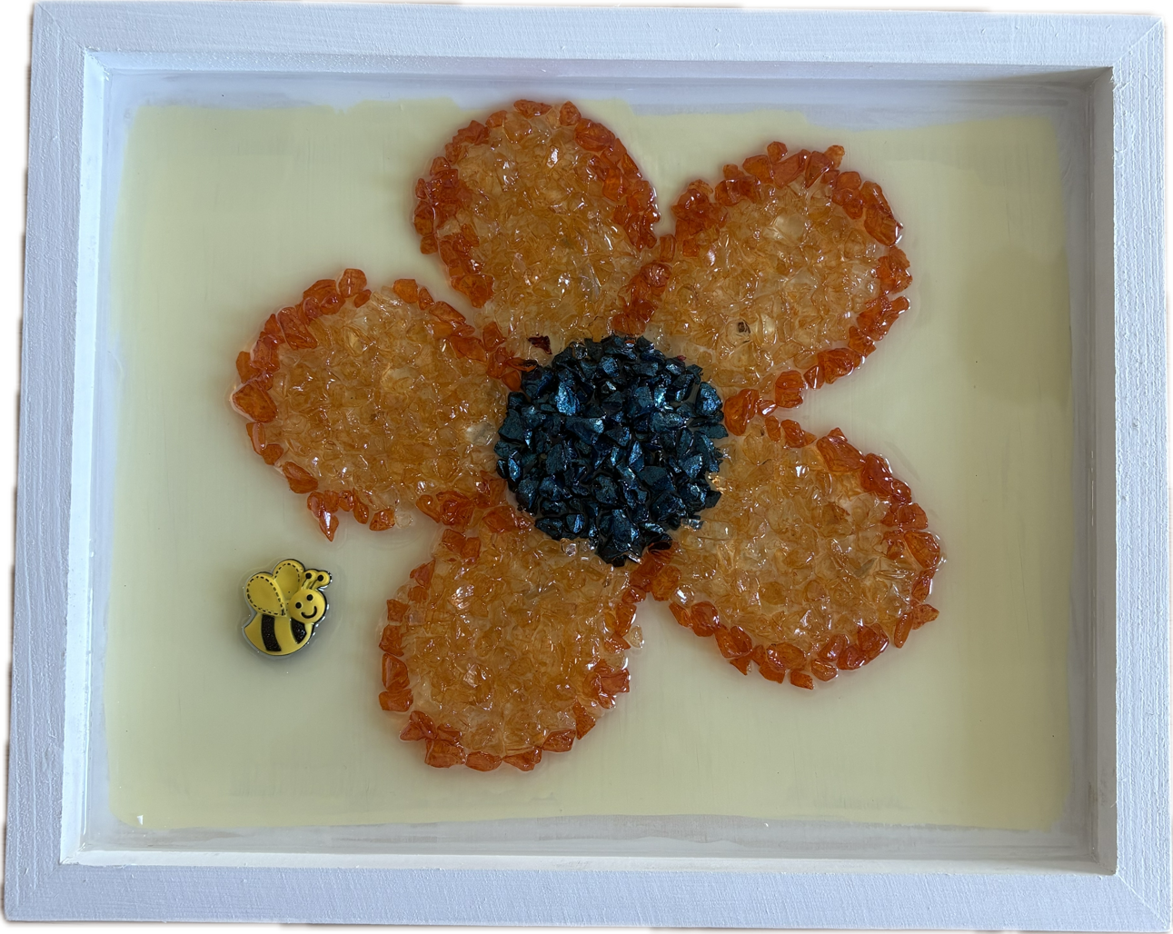 Decorative resin art of a glass flower of orange and black gemstones arranged in the shape of a flower with a bee charm at the bottom left corner. The background is a pale yellow surface within a white frame.