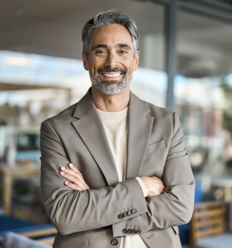 A smiling middle-aged man with gray hair and beard, wearing a light gray blazer and white shirt, standing with arms crossed in a modern office or cafe setting.