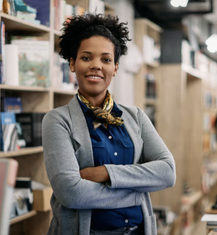 A confident young woman with curly hair standing with arms crossed in a bookstore, smiling at the camera.