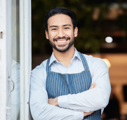 Smiling man in a light blue shirt and apron standing next to a door with arms crossed.