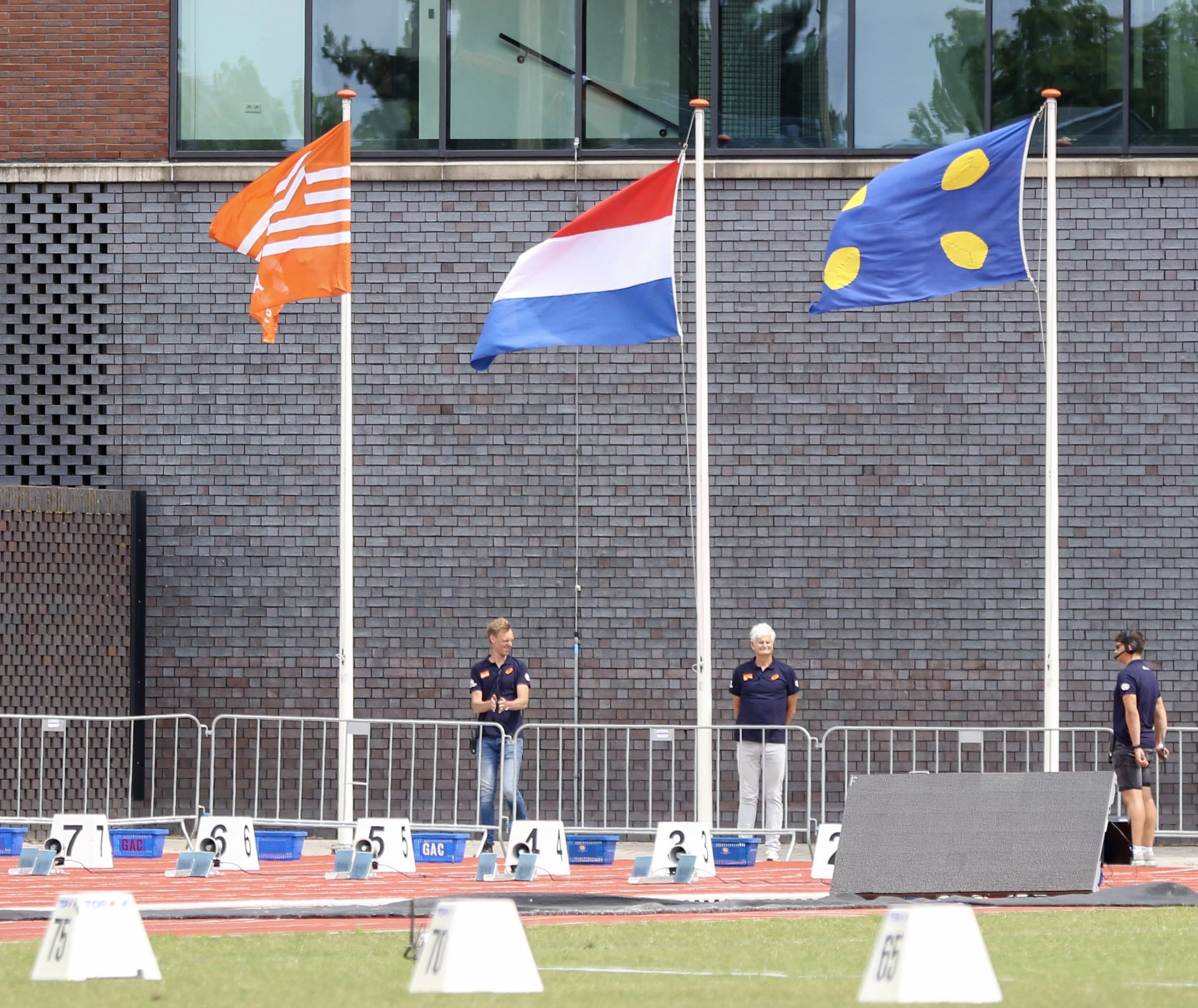 People standing in front of flags and numbered markers at an outdoor track event.