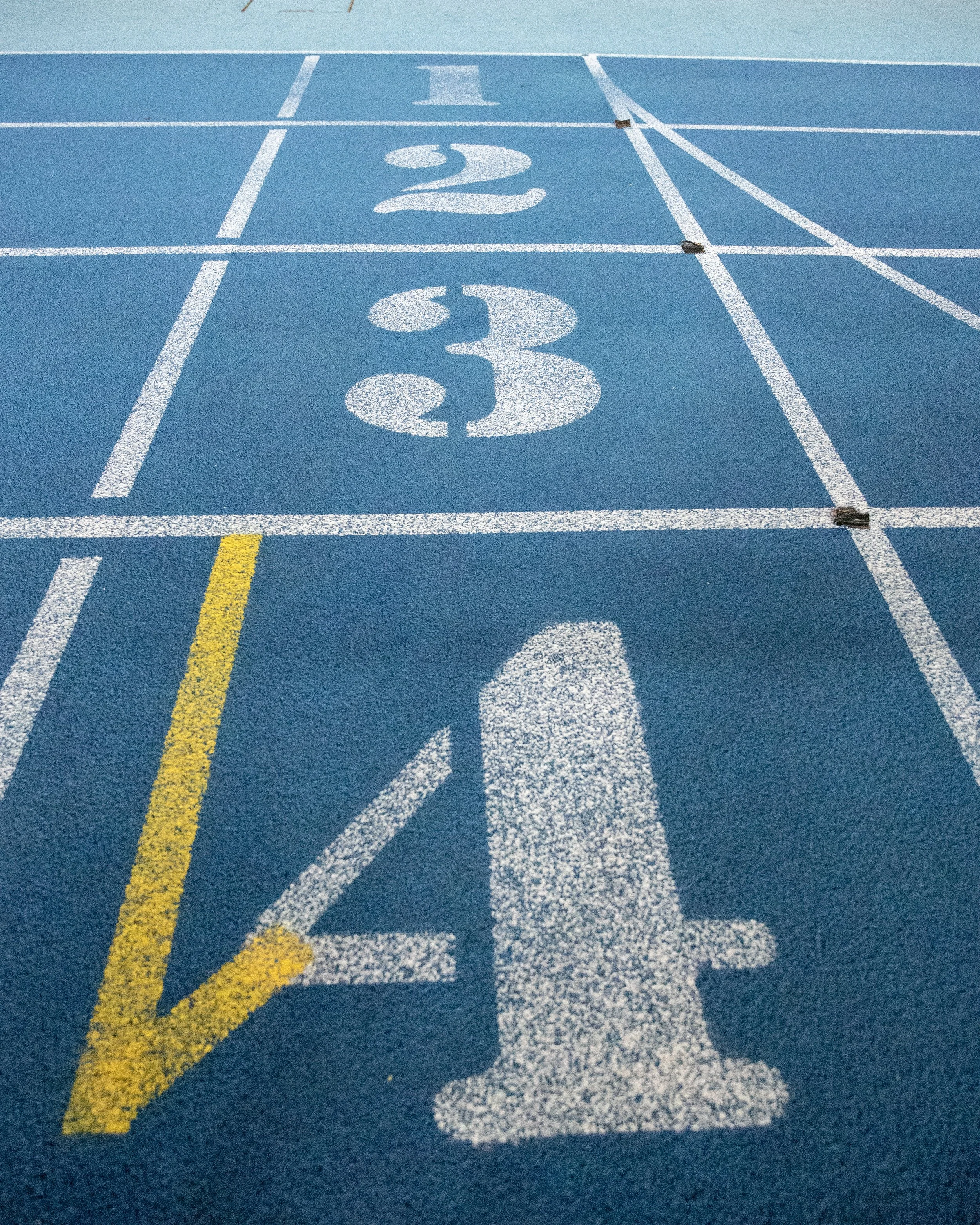 A blue running track with lanes numbered one, two, and three in white paint. Lane one is outlined in yellow. The track surface has a textured appearance.