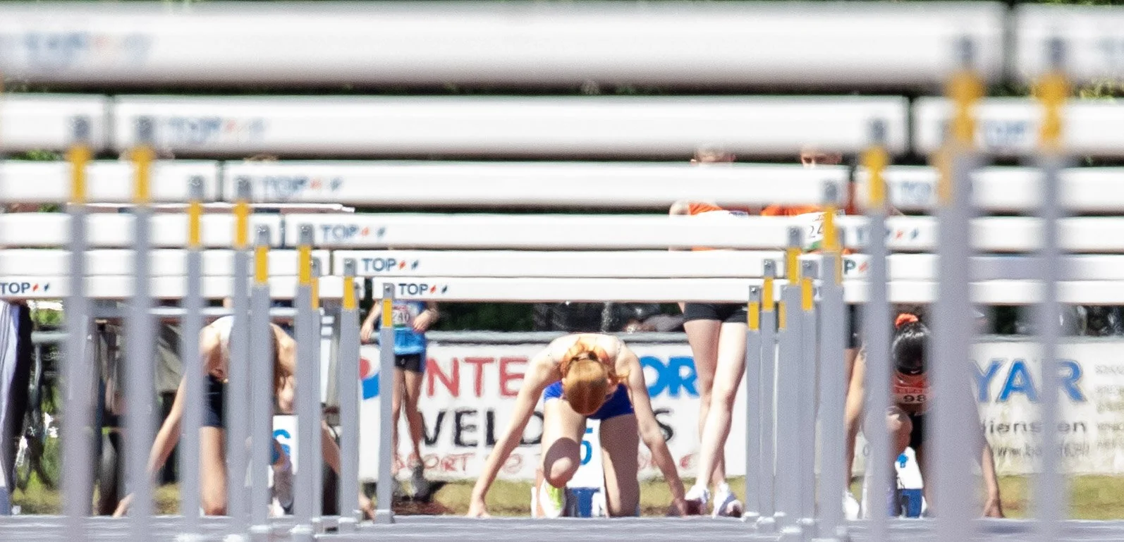 Track athletes in starting blocks preparing to race at the starting line with hurdles ahead and a banner in the background.