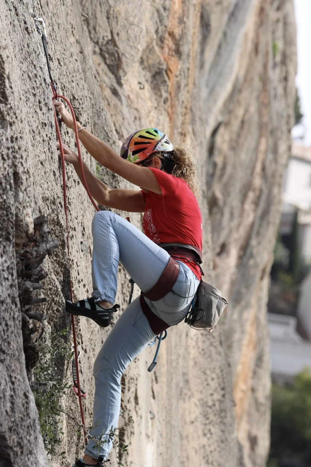 A woman with curly blond hair, wearing a red shirt, light blue jeans, climbing a rock wall with safety gear, including a colorful helmet and harness.