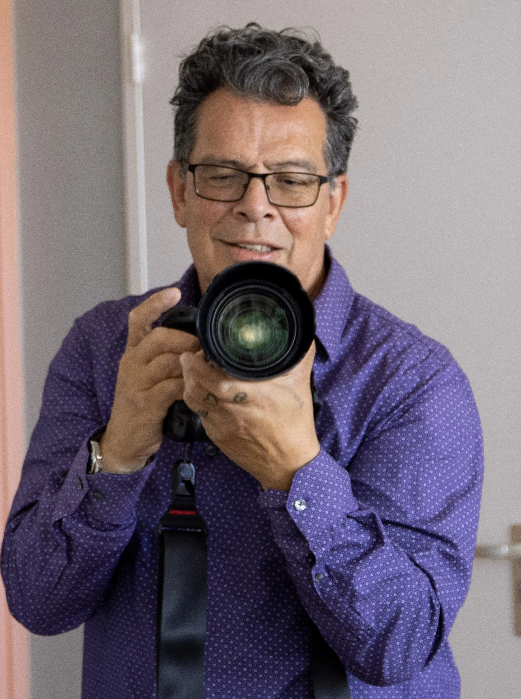 A man with curly gray hair and glasses taking a photo with a DSLR camera, wearing a purple shirt with small white dots.