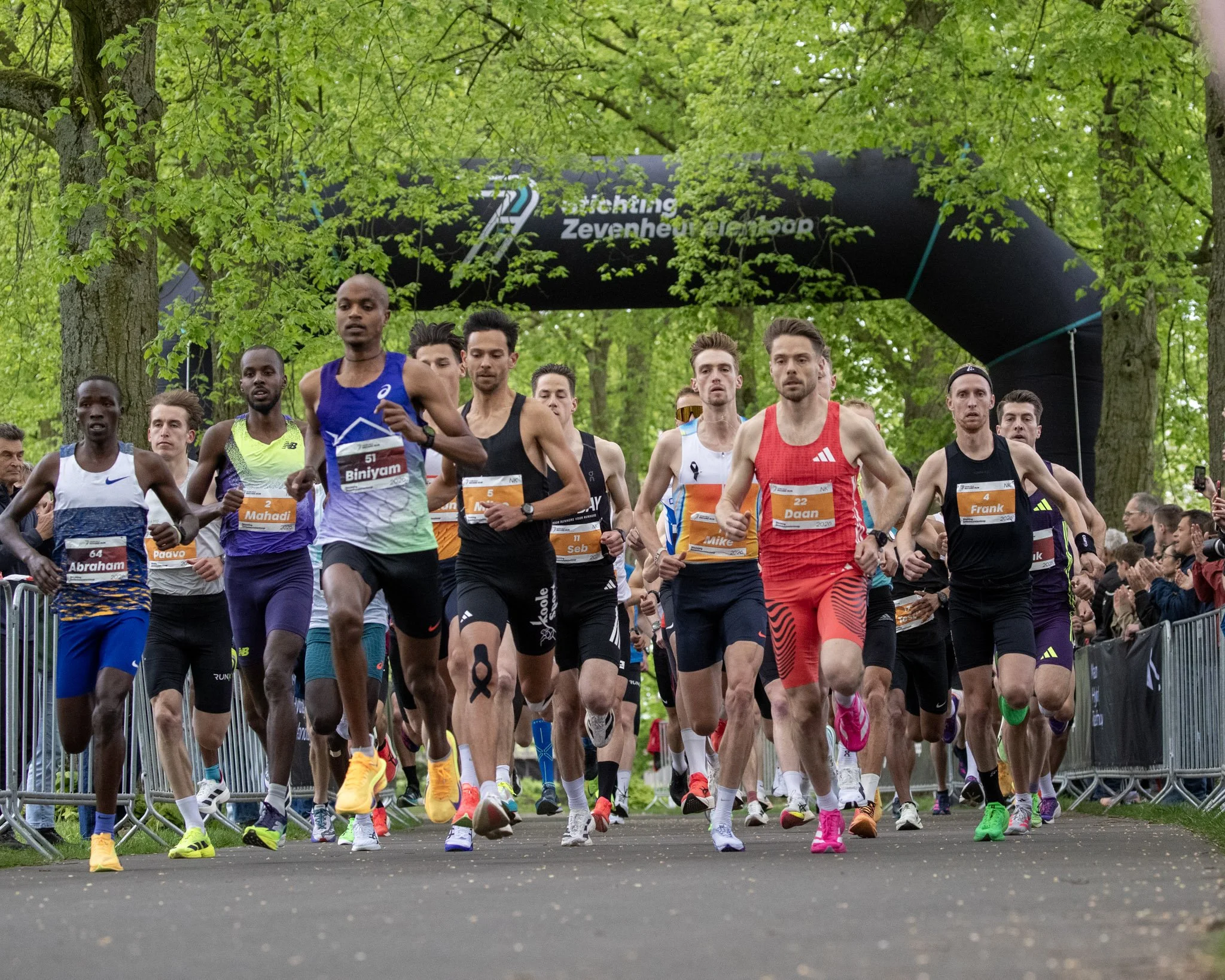 A group of marathon runners at the start of a race in a wooded park setting, with many wearing colorful athletic gear and race bibs.