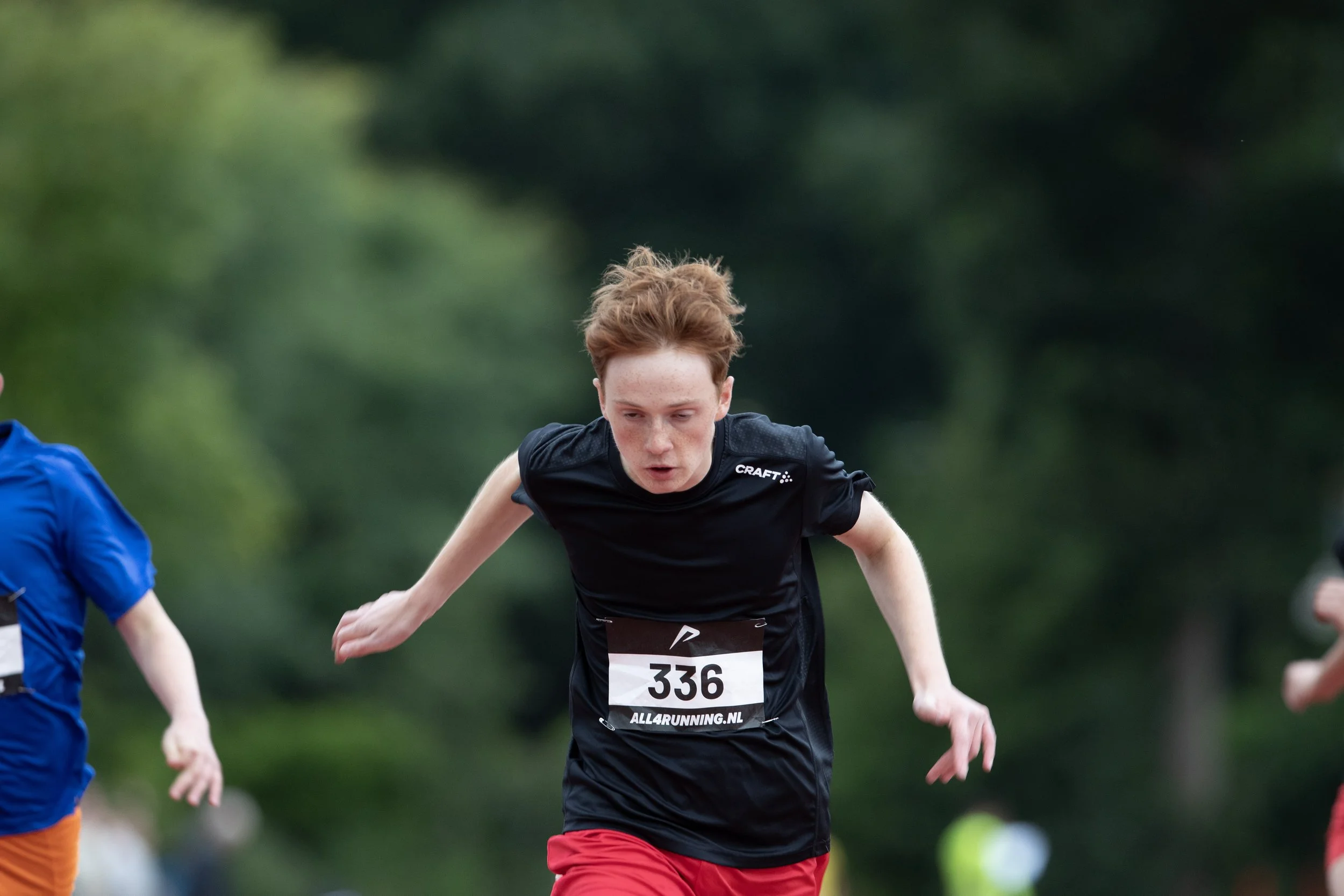 A young male runner with red hair in a black running shirt and red shorts participating in a race, with a race bib number 336, outdoors with green trees in the background.