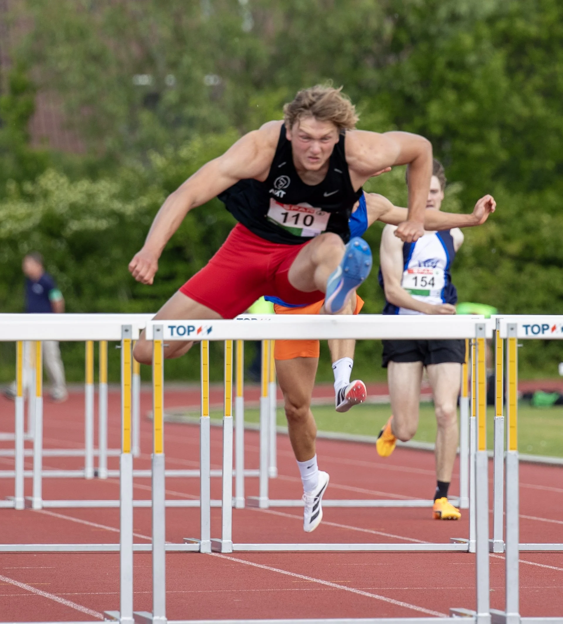 Young male athlete with curly hair in black sports top and red shorts in midair jumping over hurdles during a race on a red track field, with other competitors and green trees in the background.
