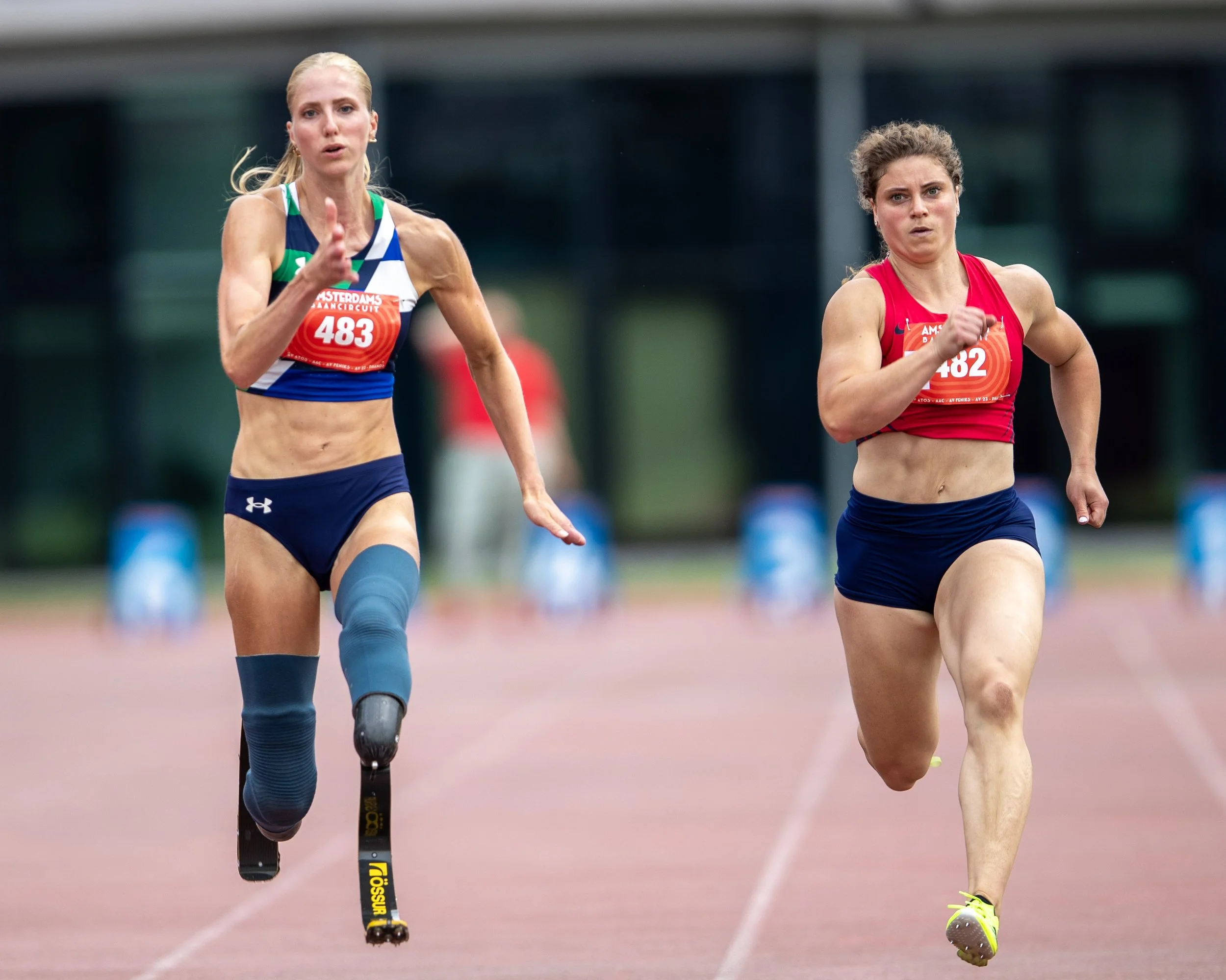 Two female runners competing in a track race, one with a prosthetic leg, on a red running track.