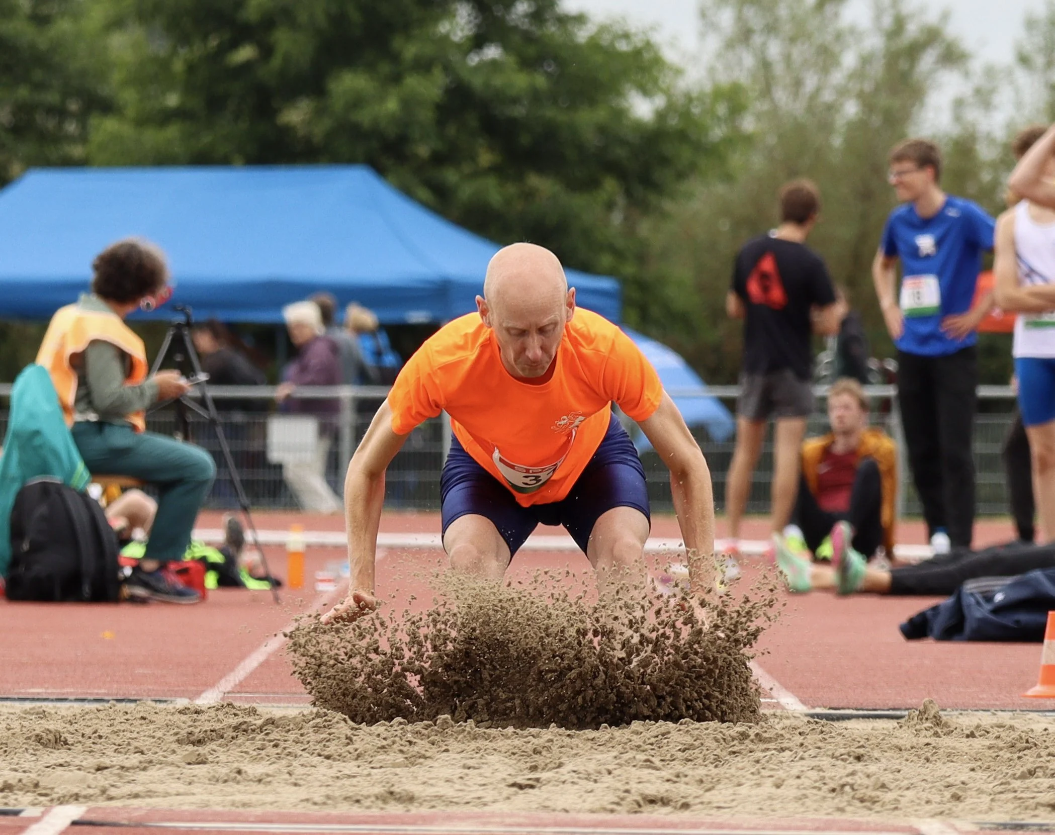 A male athlete with a shaved head wearing an orange shirt and dark shorts performing a long jump onto sand at an outdoor track and field event. Spectators and other athletes are in the background.