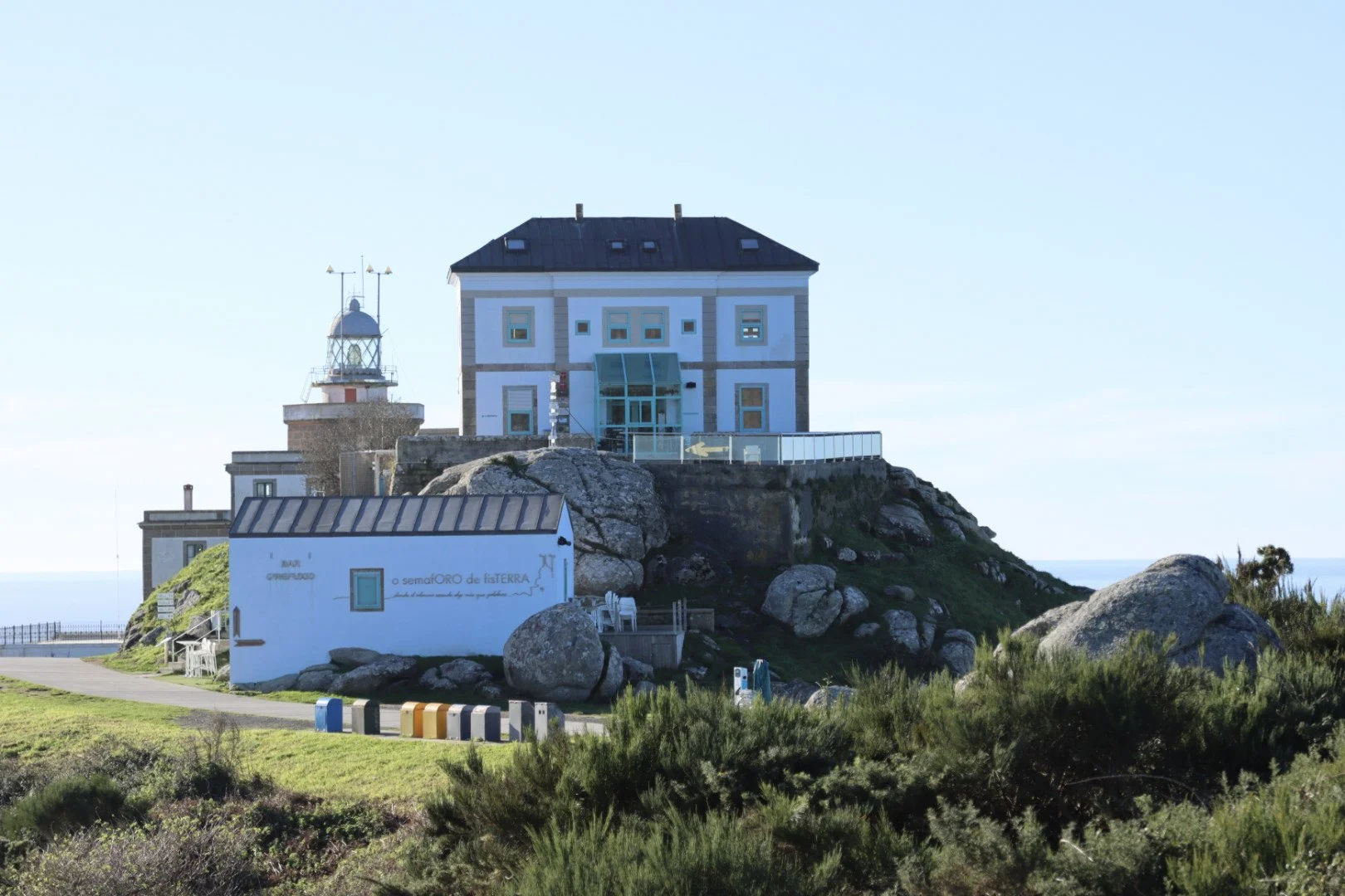 A lighthouse and a large building on a rocky hill, with green bushes in the foreground and a clear blue sky in the background.