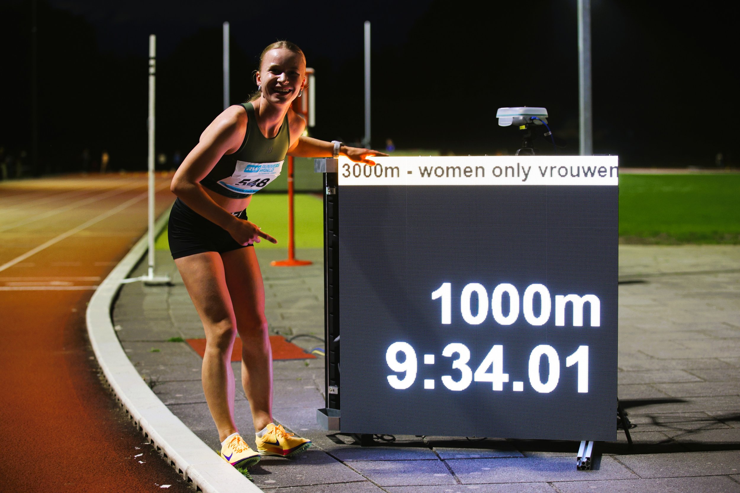 A female runner smiling and pointing at a digital timer at a nighttime track event. The timer displays completed distances of 1000 meters, 2000 meters, and 3000 meters along with times. The woman is wearing a sports bra, shorts, running shoes, and a watch, and she is standing near a track with flags and field equipment in the background.