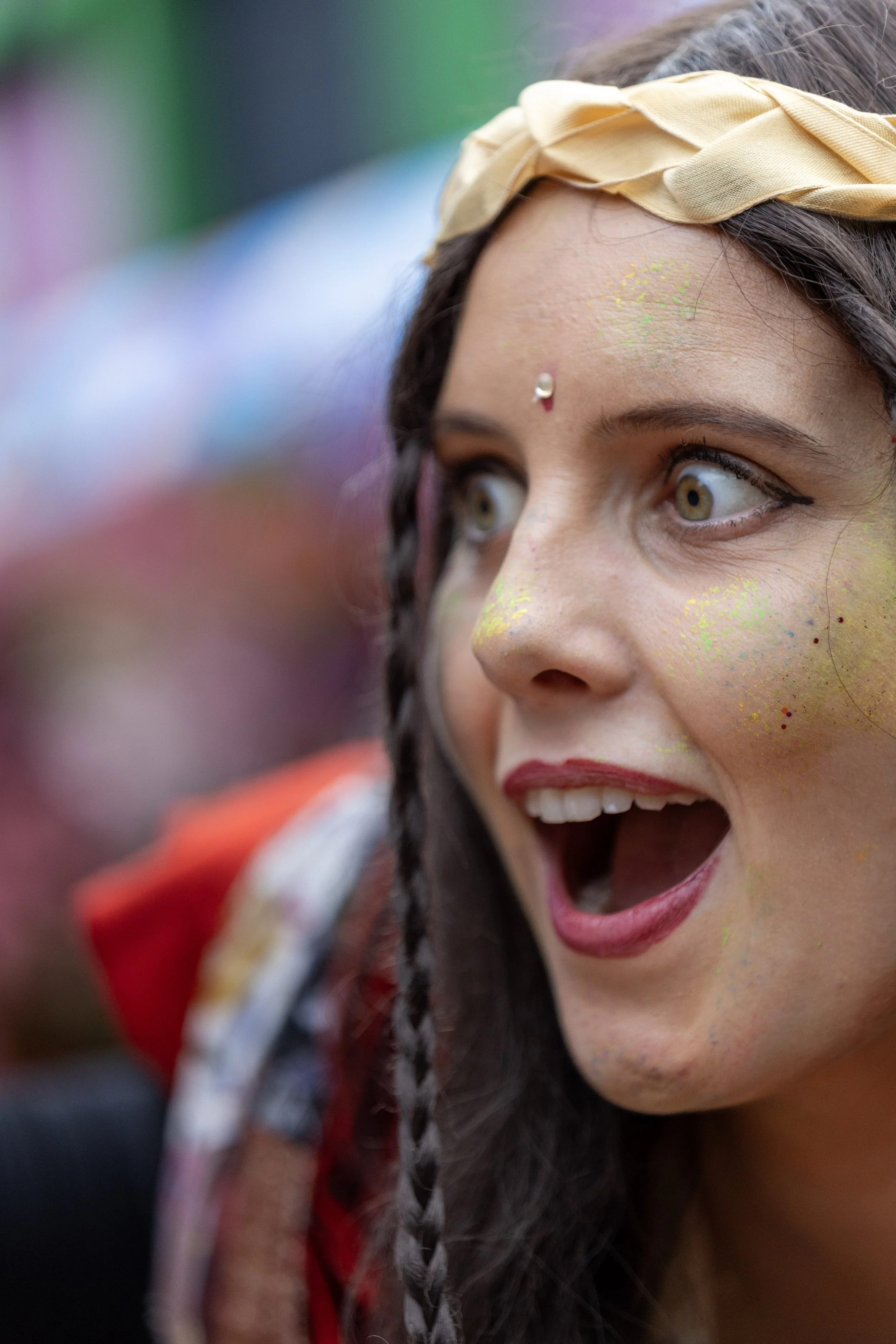 Close-up of a woman with painted face, wearing a beige headband, with green and yellow paint on her face, and her mouth open as if shouting or cheering.