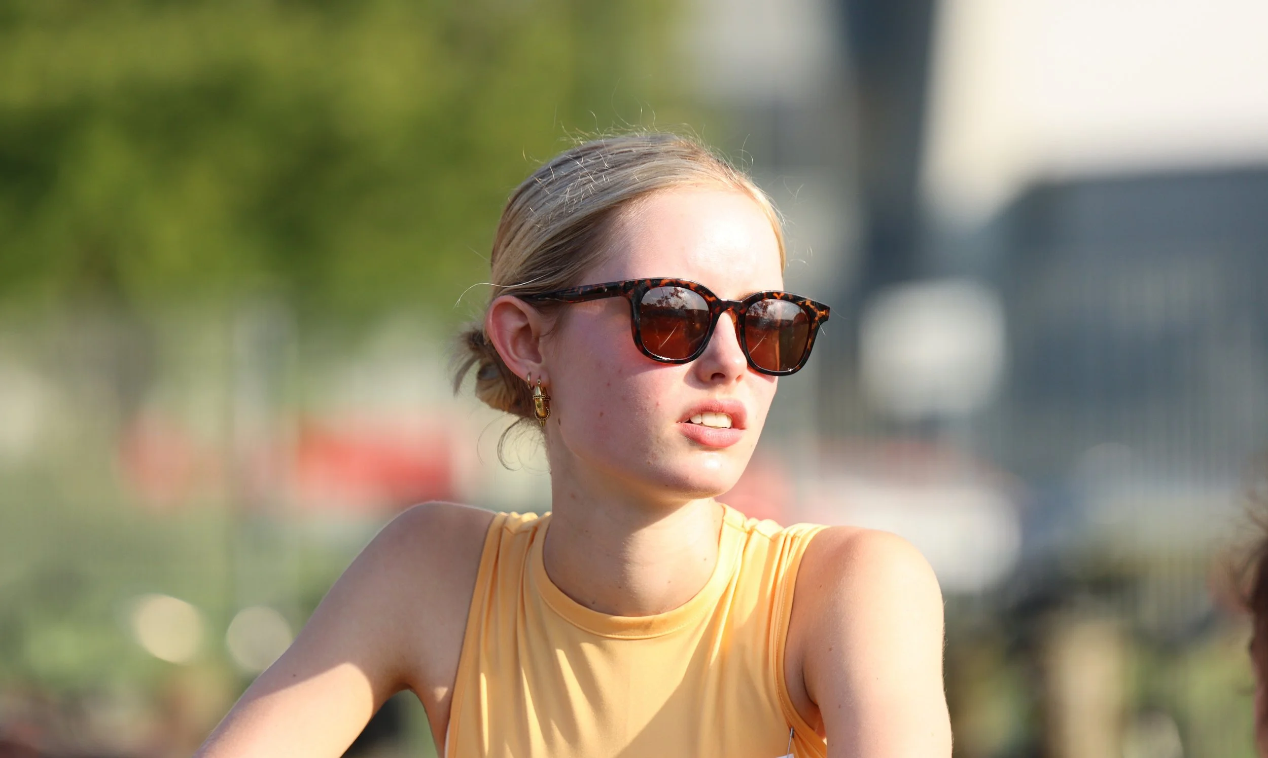 Young woman with blonde hair wearing tortoiseshell sunglasses and a yellow sleeveless top outdoors.