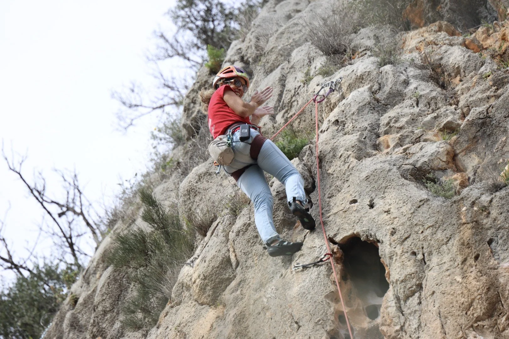 Woman in red shirt and helmet rock climbing on a vertical rock face with climbing gear and safety rope.