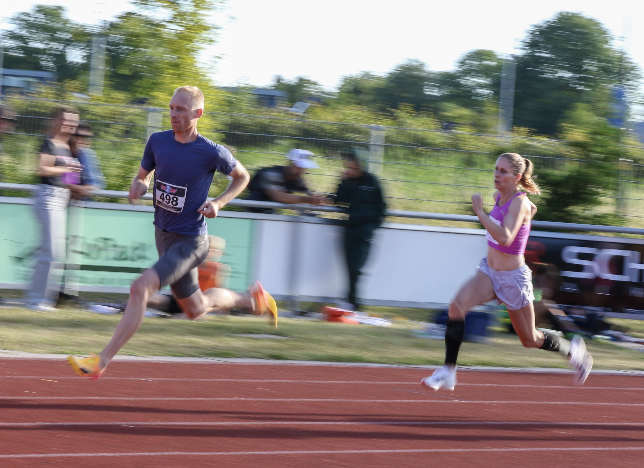 Two athletes running on a track during a race, with spectators and greenery in the background.