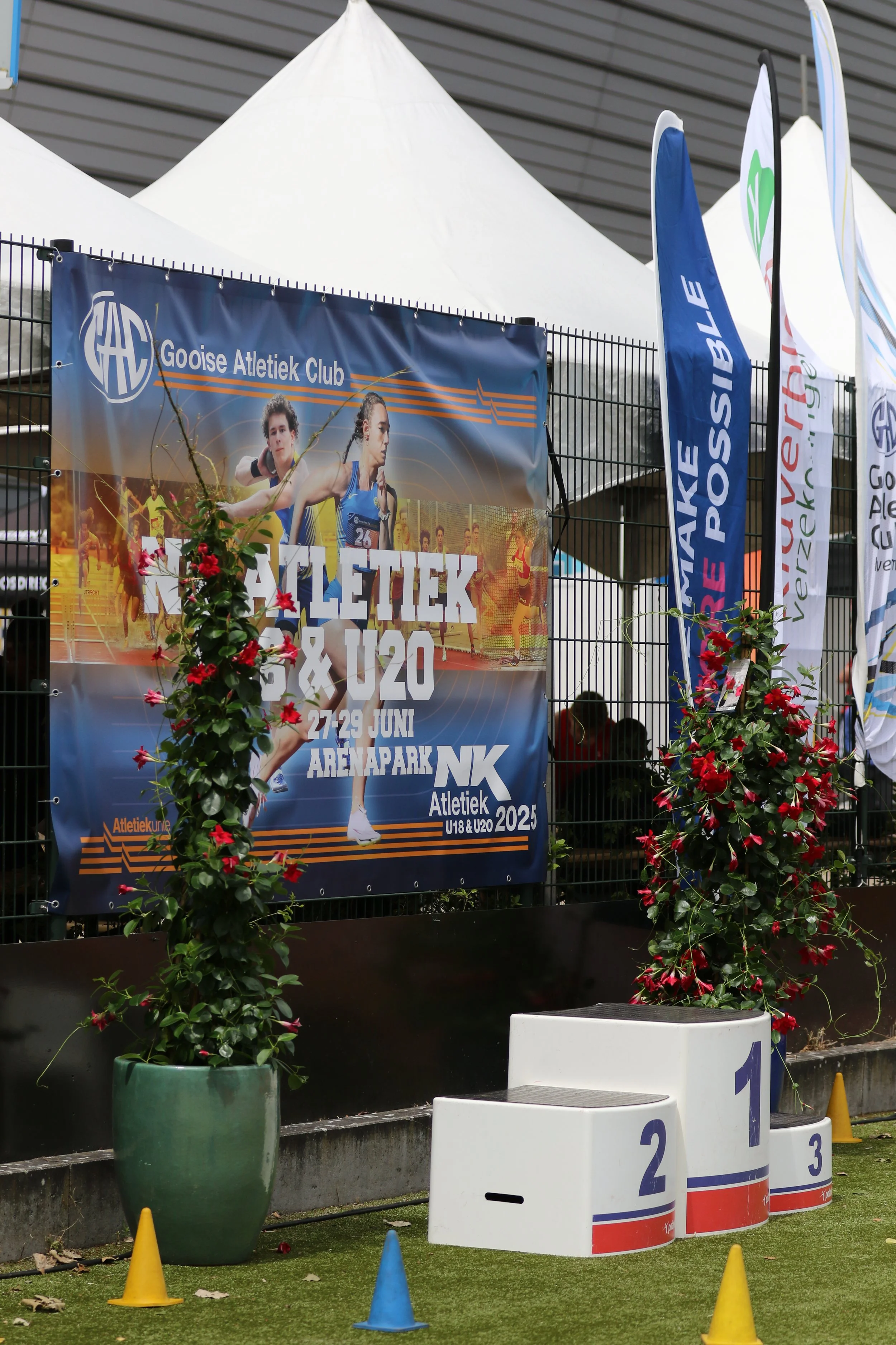 A podium for awards with spaces for first, second, and third place, decorated with two large potted plants with red flowers, and several small colored cones on artificial grass. Behind, there is a banner advertising a track and field event with images of athletes, and some flags.