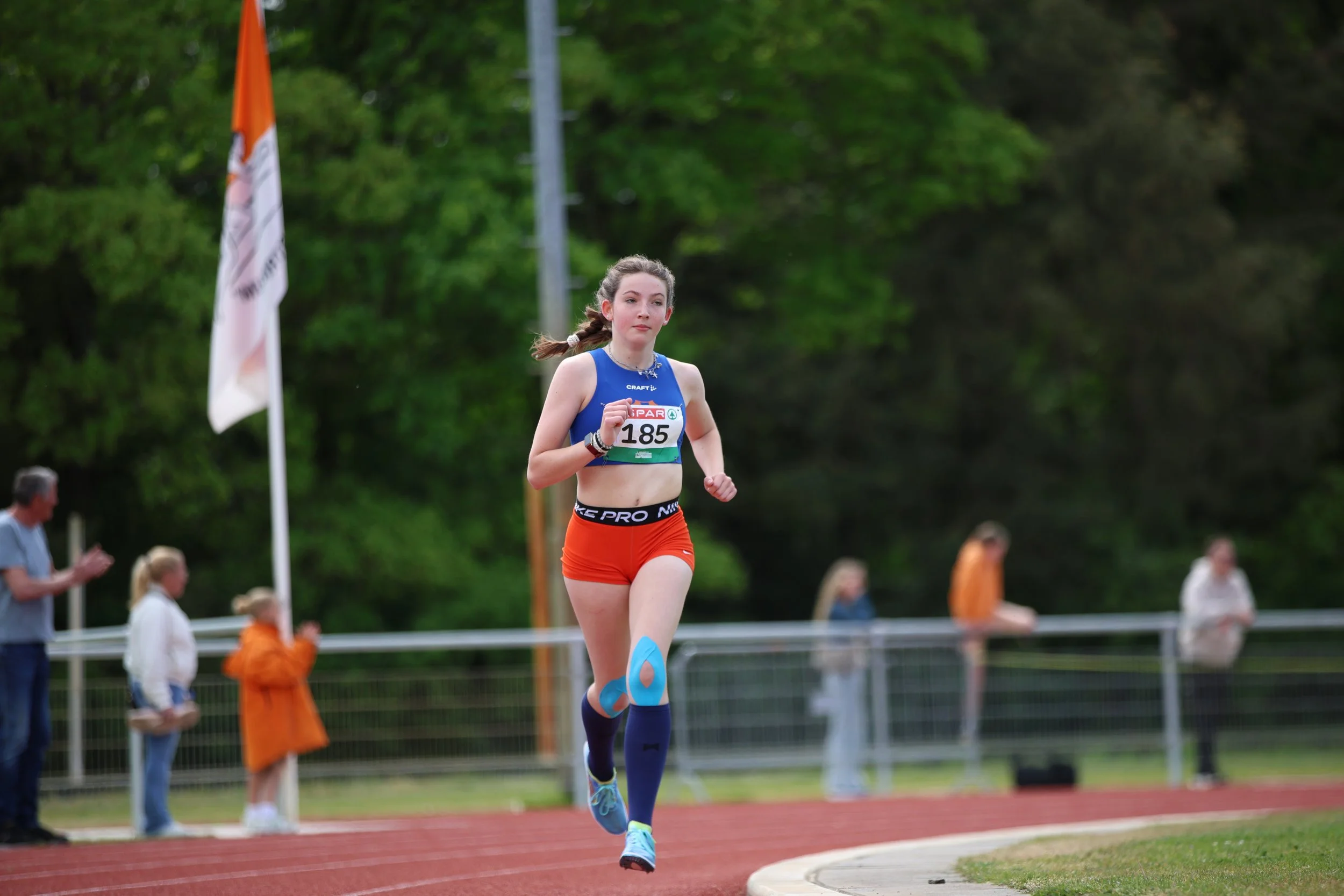 A young female athlete running on a track during a race, surrounded by spectators and volunteers, with trees in the background.