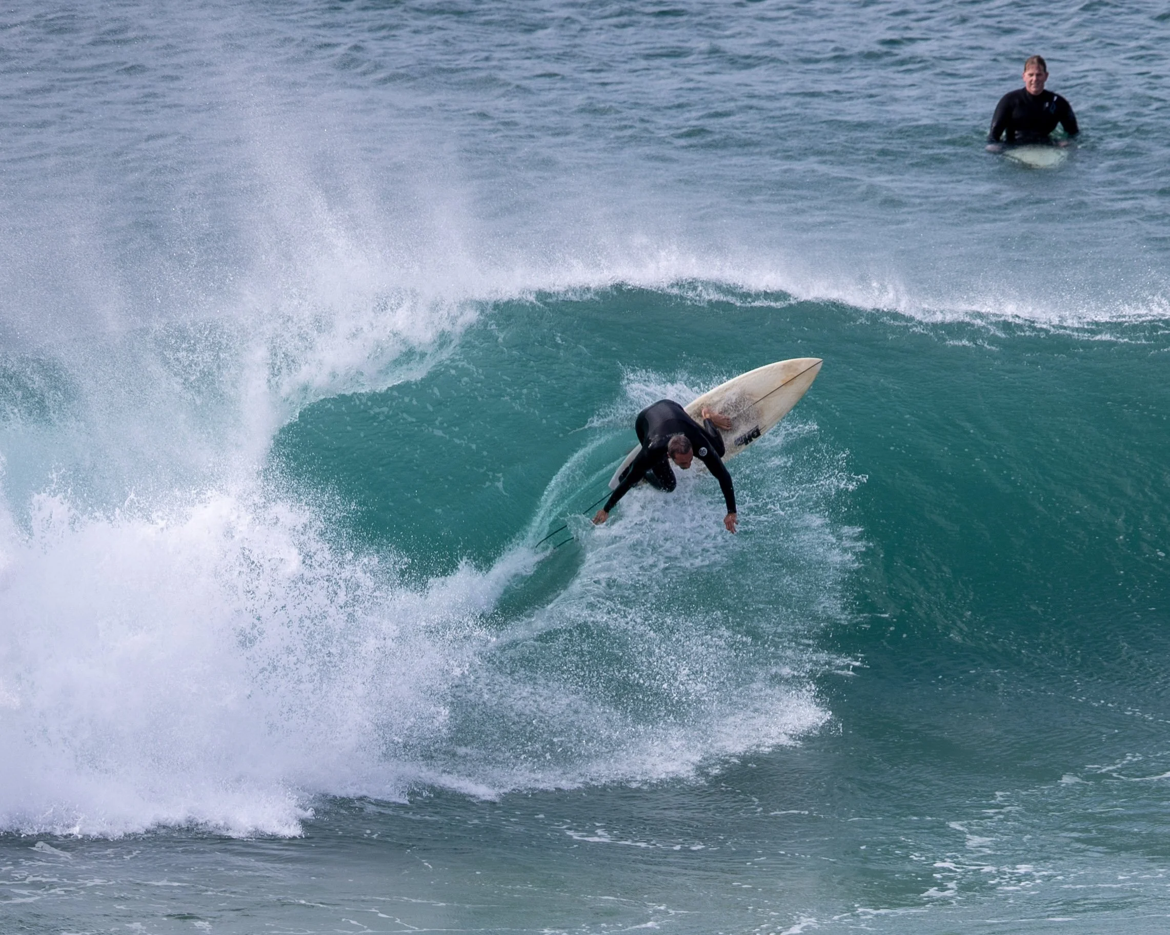 A person in a wetsuit surfing on a wave with another person in the background standing in the water.