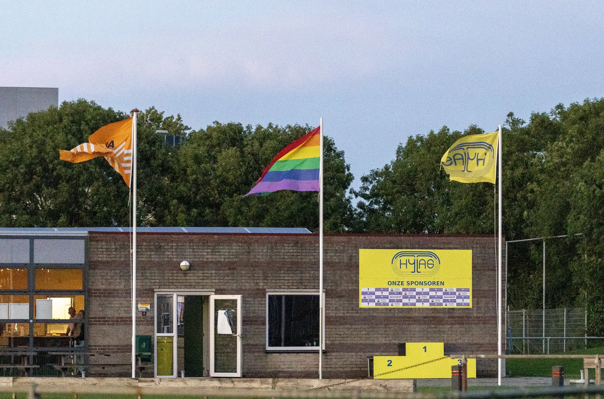 Three flags flying outside a building with a brick wall, including a rainbow pride flag, an orange flag, and a yellow flag, with trees in the background and a large yellow sponsor board.