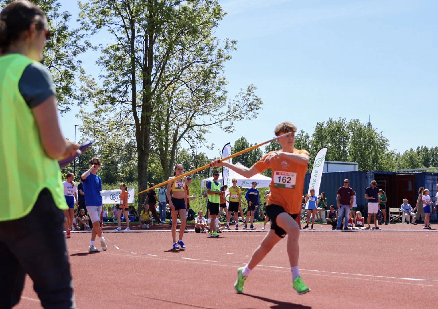 Young male athlete in orange shirt and black shorts throws a javelin on a track field during a sporting event. Spectators and other athletes watch in the background on a sunny day with trees and tents.