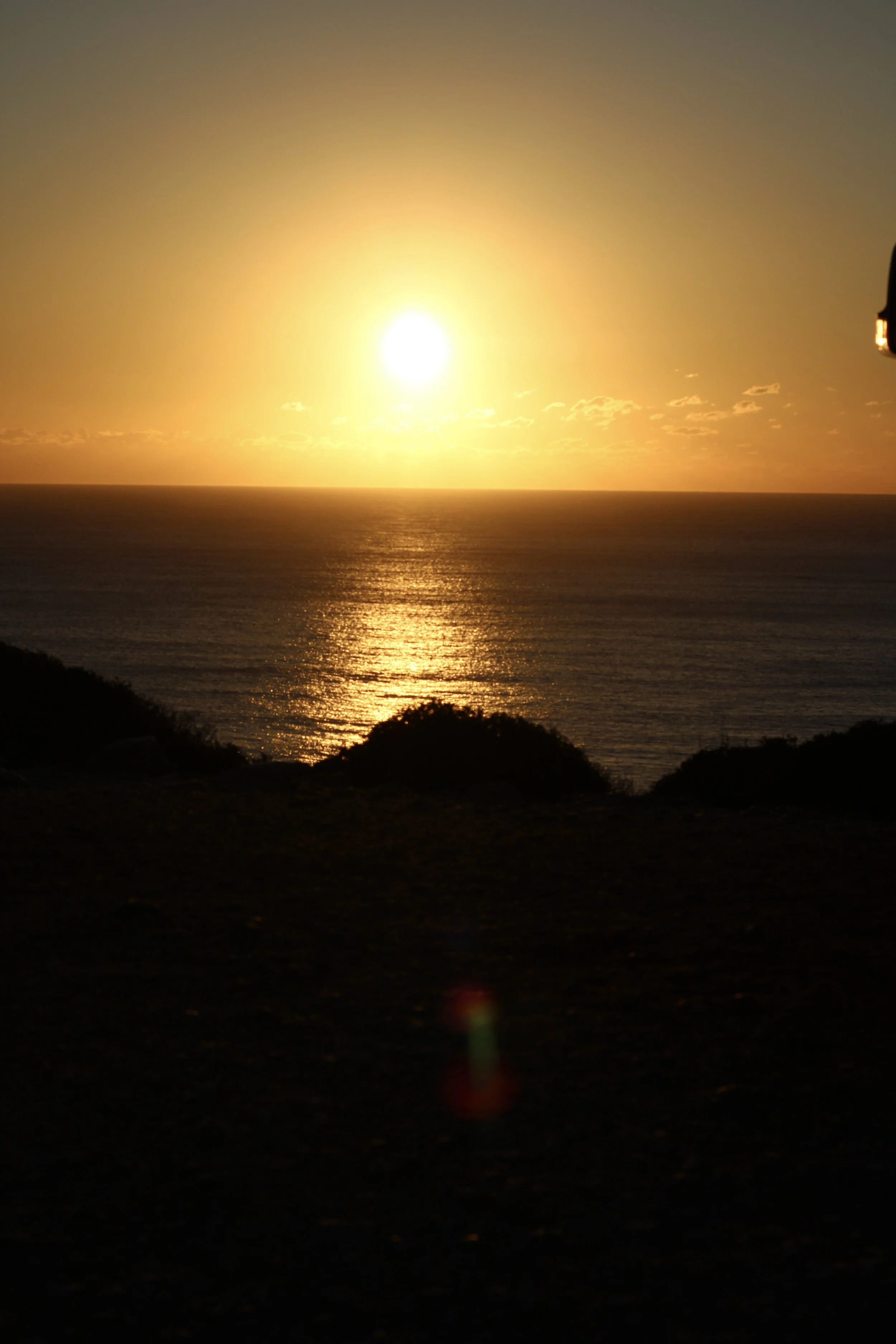 Sunset over the ocean with reflections on water, silhouetted hills in foreground, partly cloudy sky