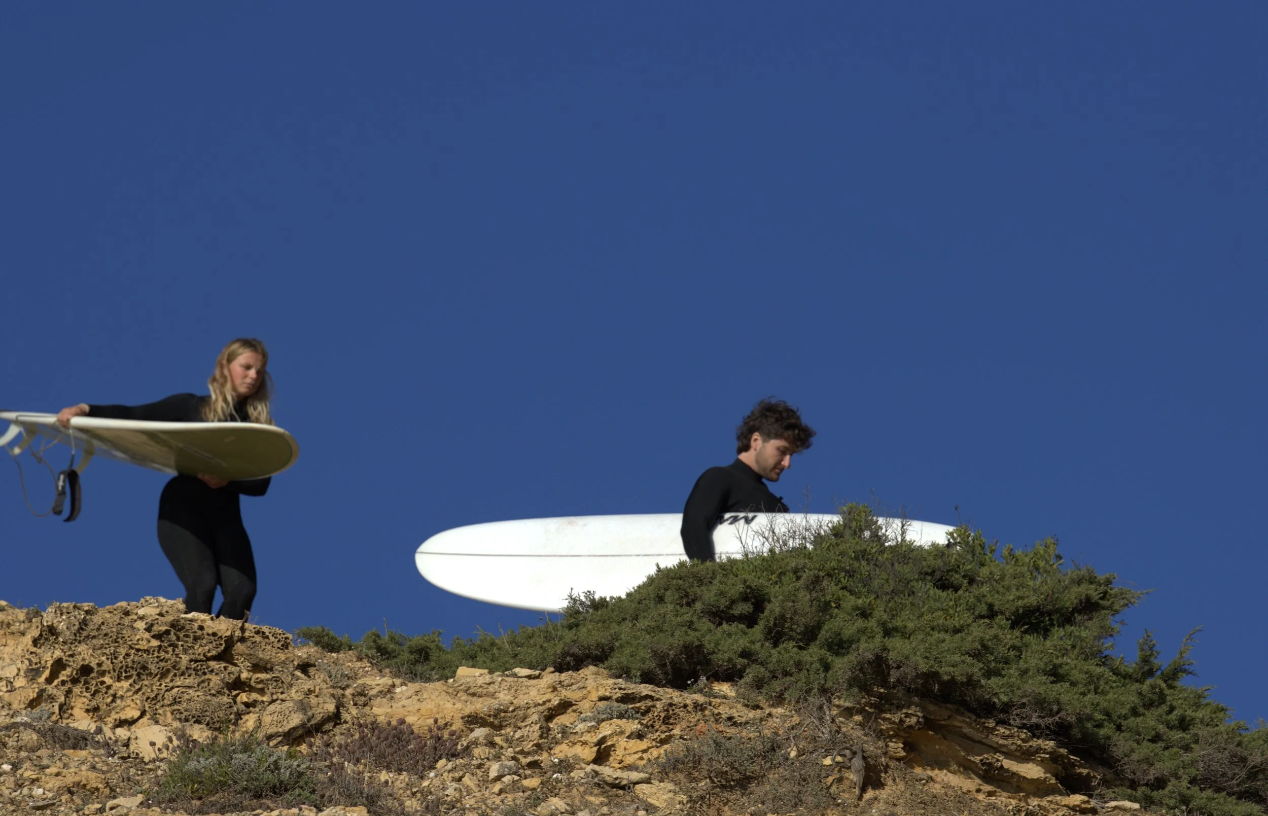 Two surfers, a woman and a man, carrying surfboards on rocky terrain against a clear blue sky.
