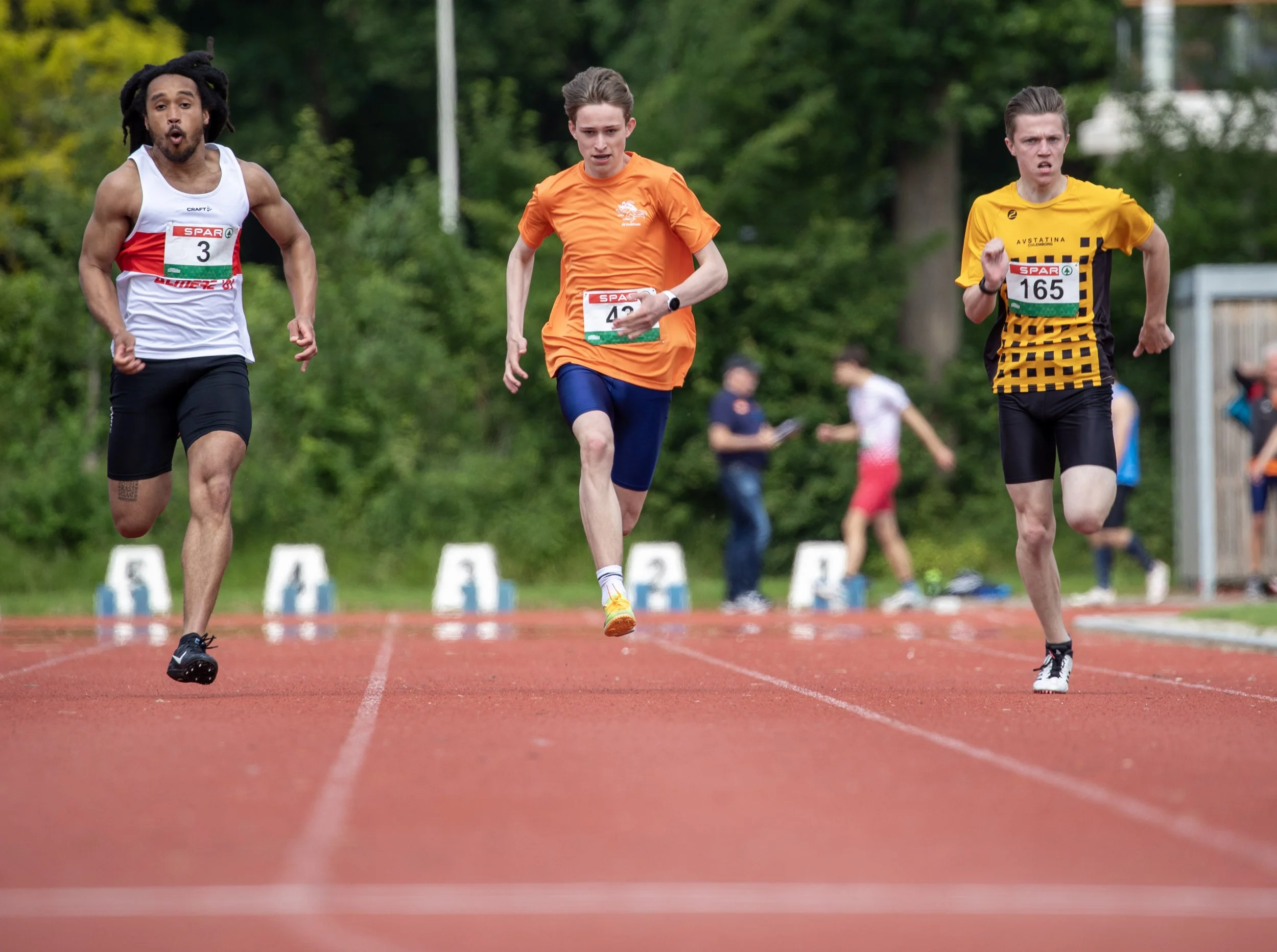 Three male runners competing in a race on a red running track with trees and spectators in the background.