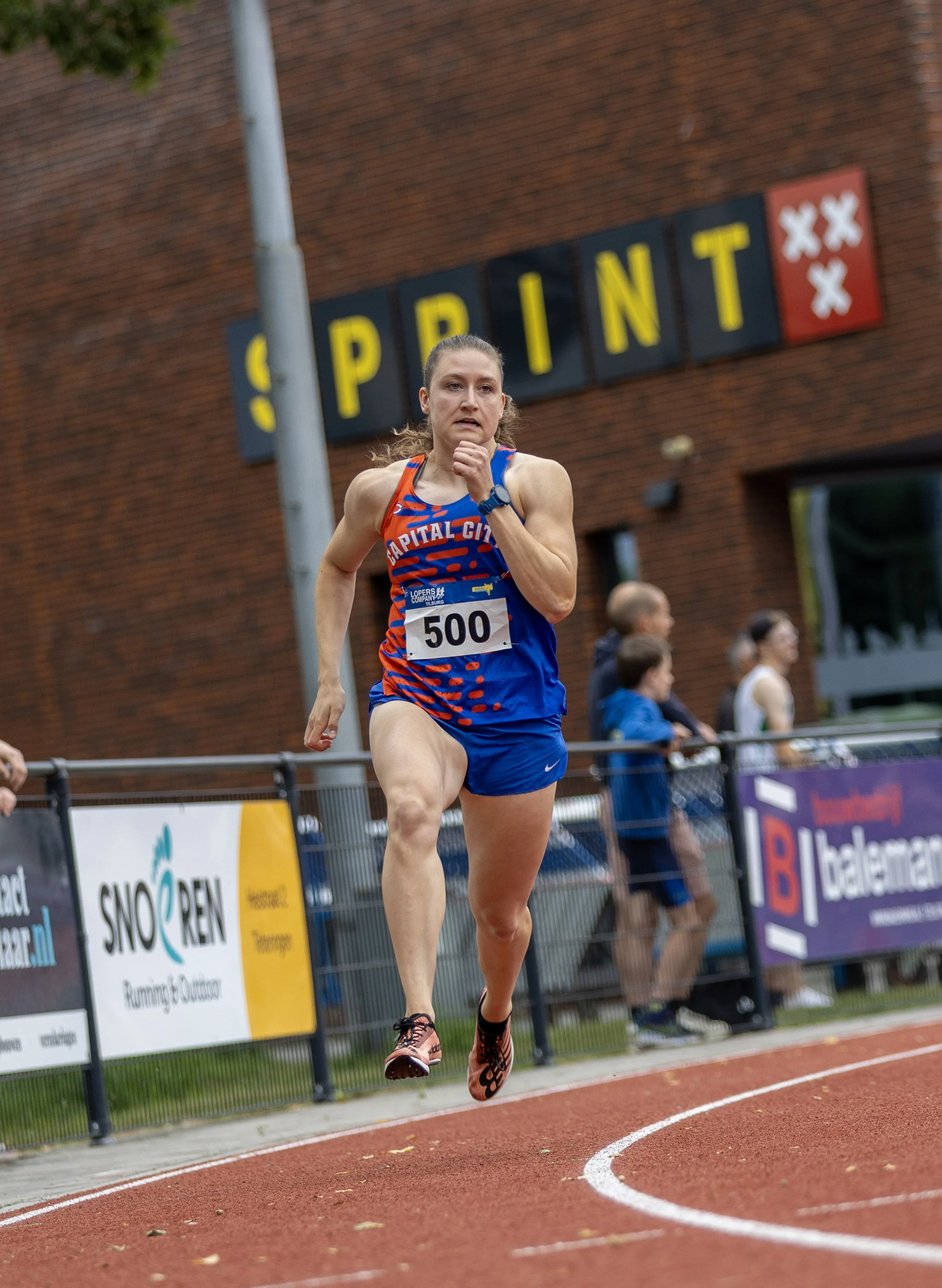 Female athlete running on a track during a race, wearing a blue and orange uniform with the number 500, in front of a brick building with a sign that says 'PRINT' and spectators watching.