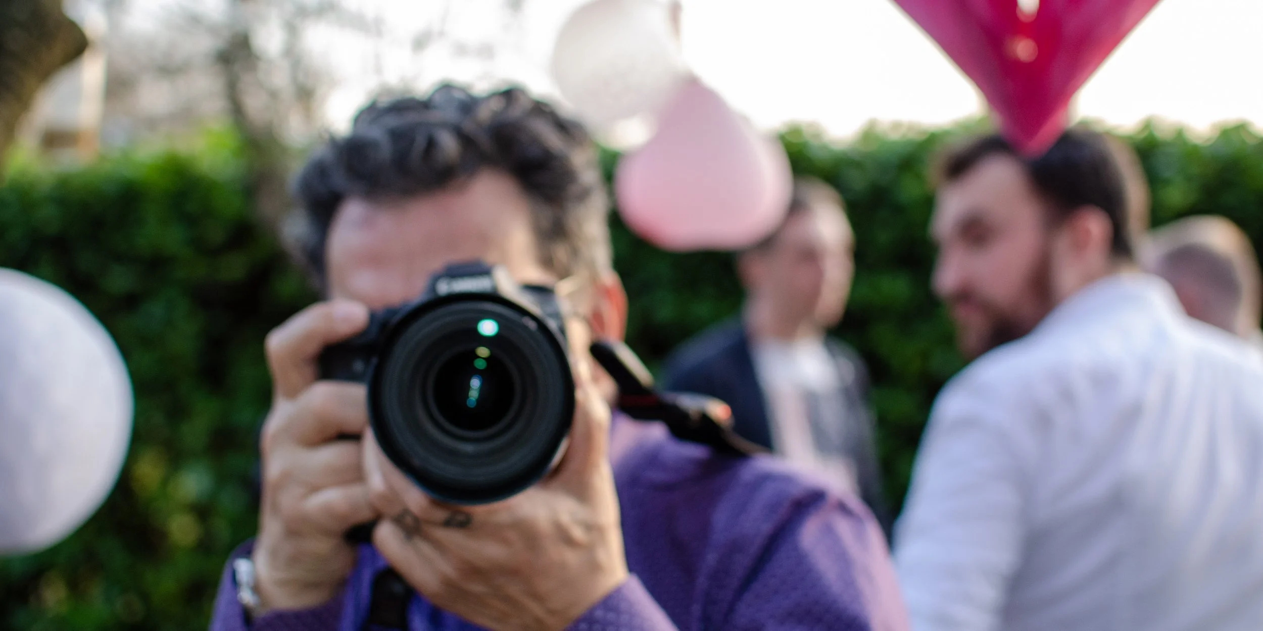 Person holding a camera taking a photo at an outdoor gathering with blurred people and pink and white balloons in the background.