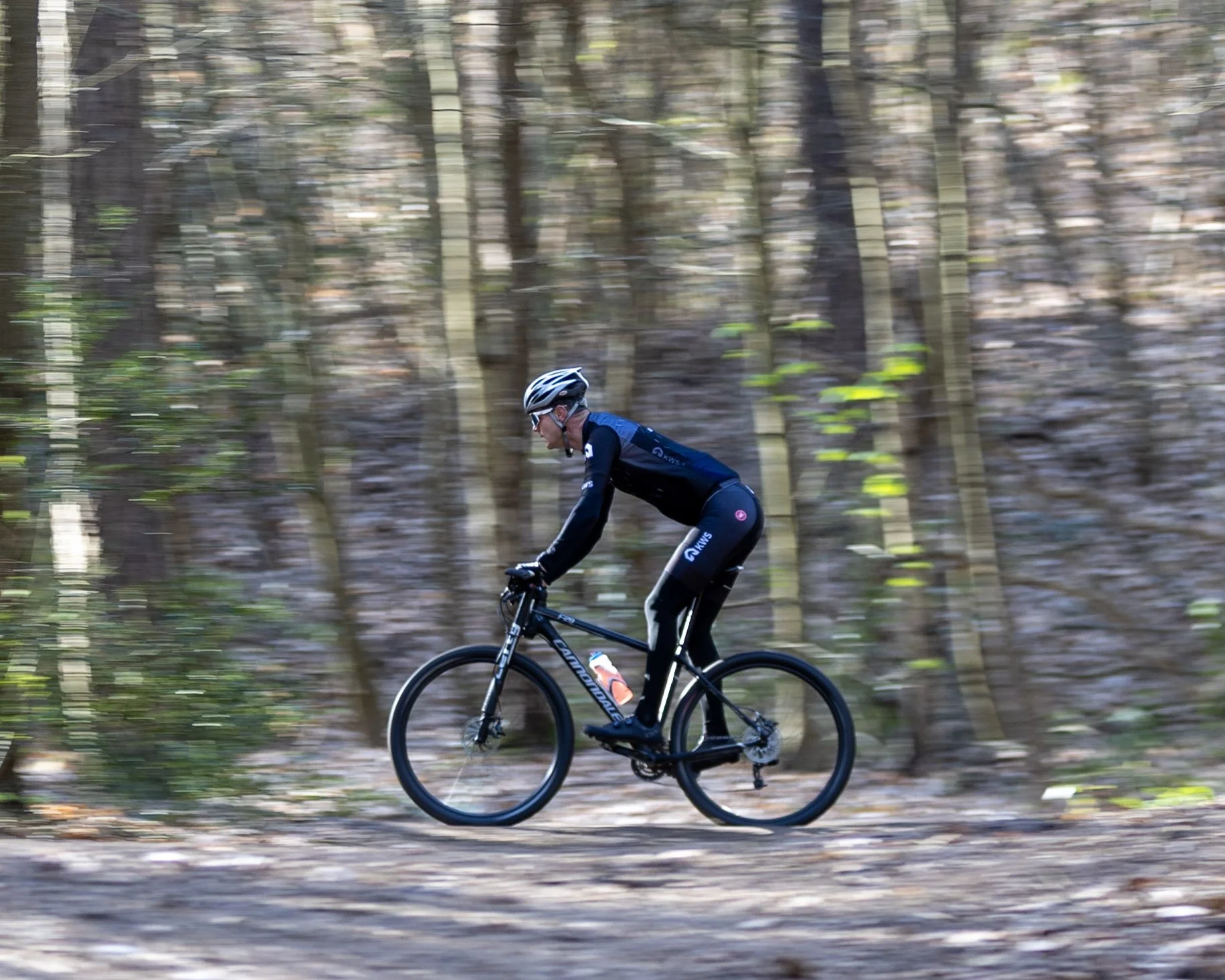 A person wearing a helmet and sunglasses riding a mountain bike through a wooded area with trees and fallen leaves.