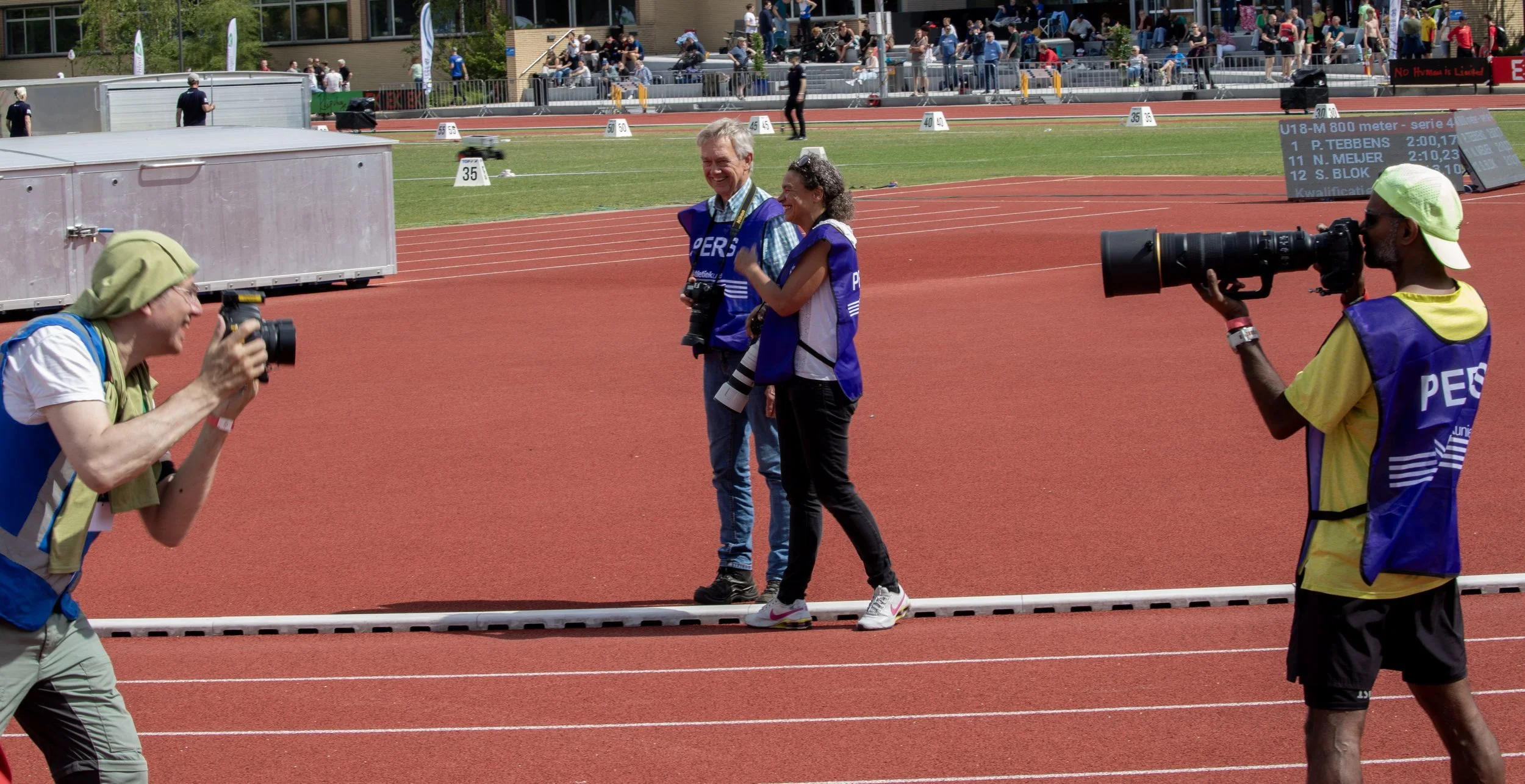 Two photographers are taking pictures of a smiling man and woman standing on a running track at a sports stadium. The man and woman are wearing blue vests, and the woman is waving or gesturing at the photographer. The background shows a stadium filled with spectators and event signage.