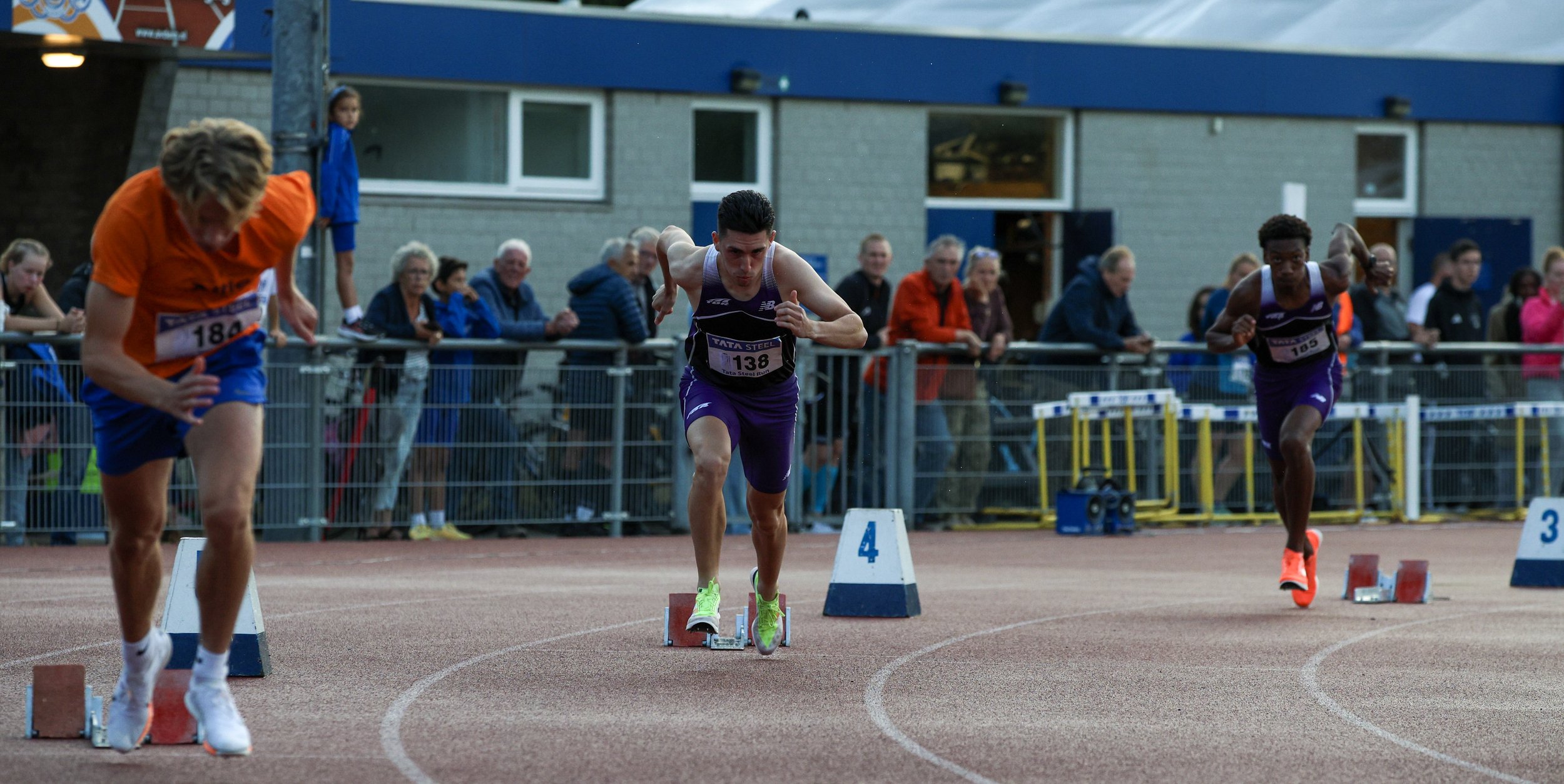 Three male athletes race in a track and field event, starting from blocks on a running track, with spectators watching behind a fence.