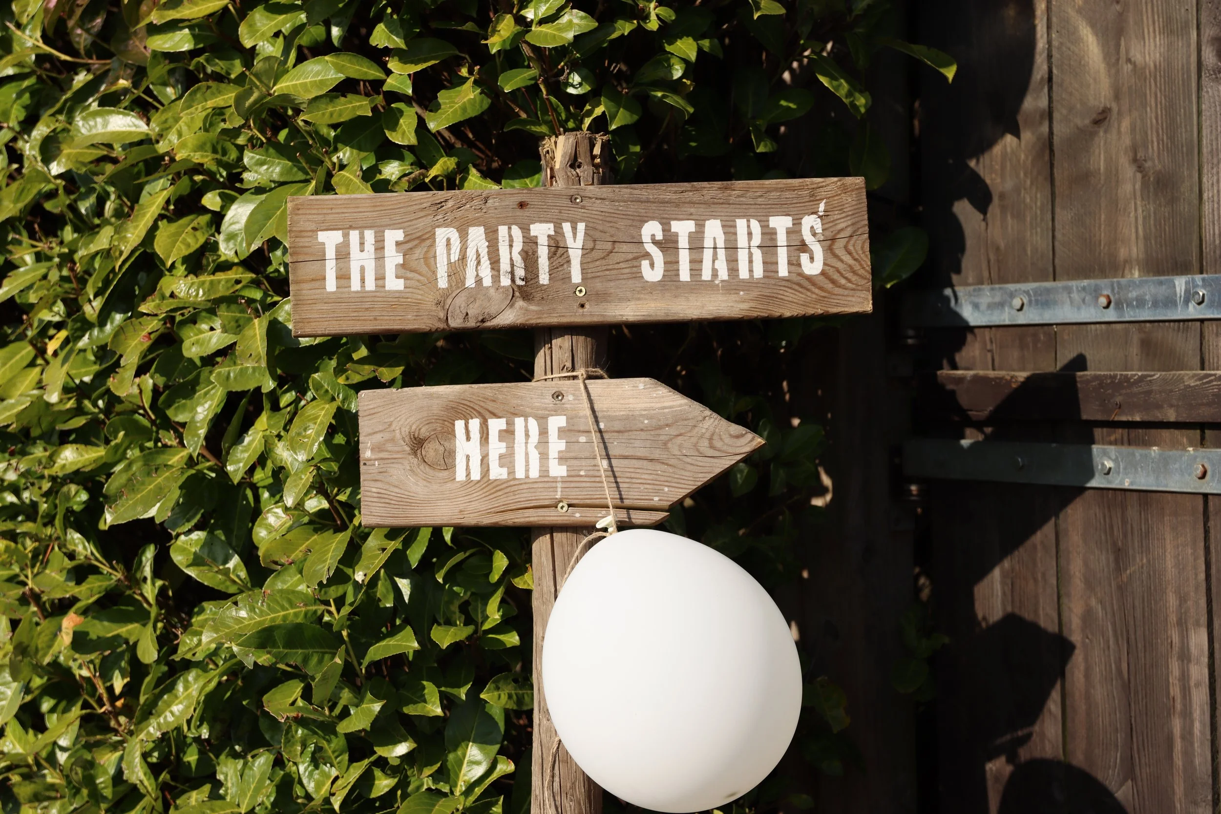 Wooden sign with white painted text that says "The party starts here" placed among green bushes and a wooden deck, with a white balloon partially visible at the bottom.