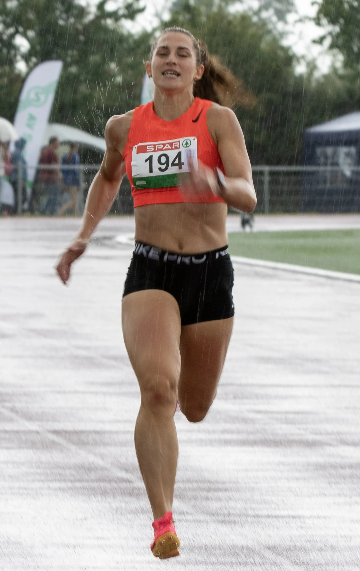 Female athlete running in a race, wearing a red sports top, black shorts, and pink shoes, with a bib number 194, in rainy weather on a track.
