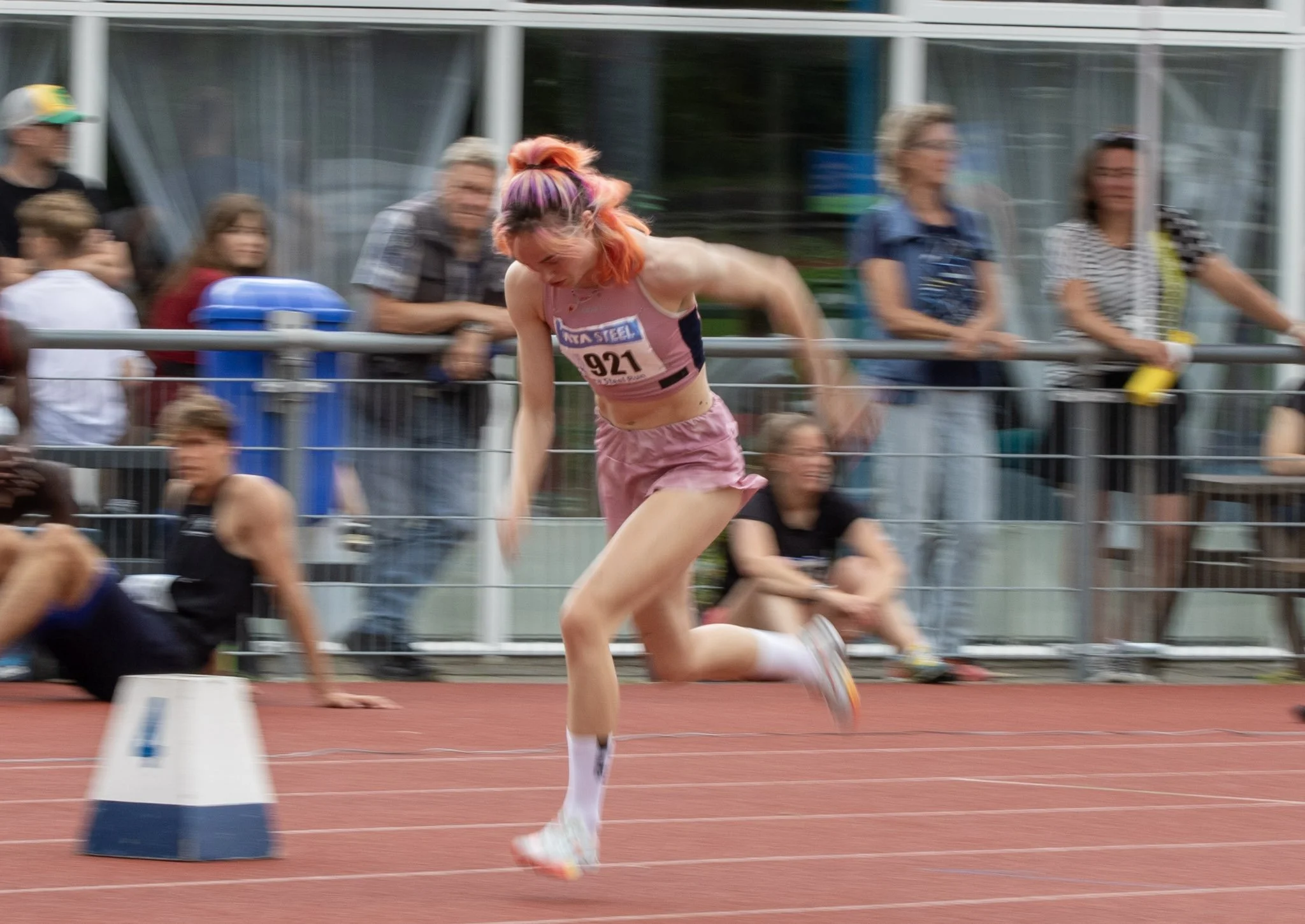A female athlete with pink hair in a pink sports bra and shorts crosses the finish line in a track race at an outdoor stadium, with spectators watching behind a fence.