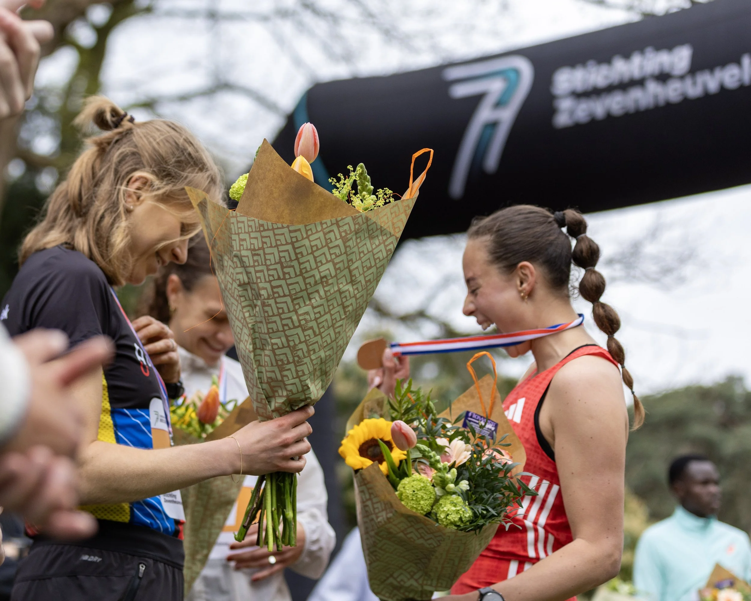 Female athletes holding bouquets and medals after a race, with medals around their necks, outdoors.