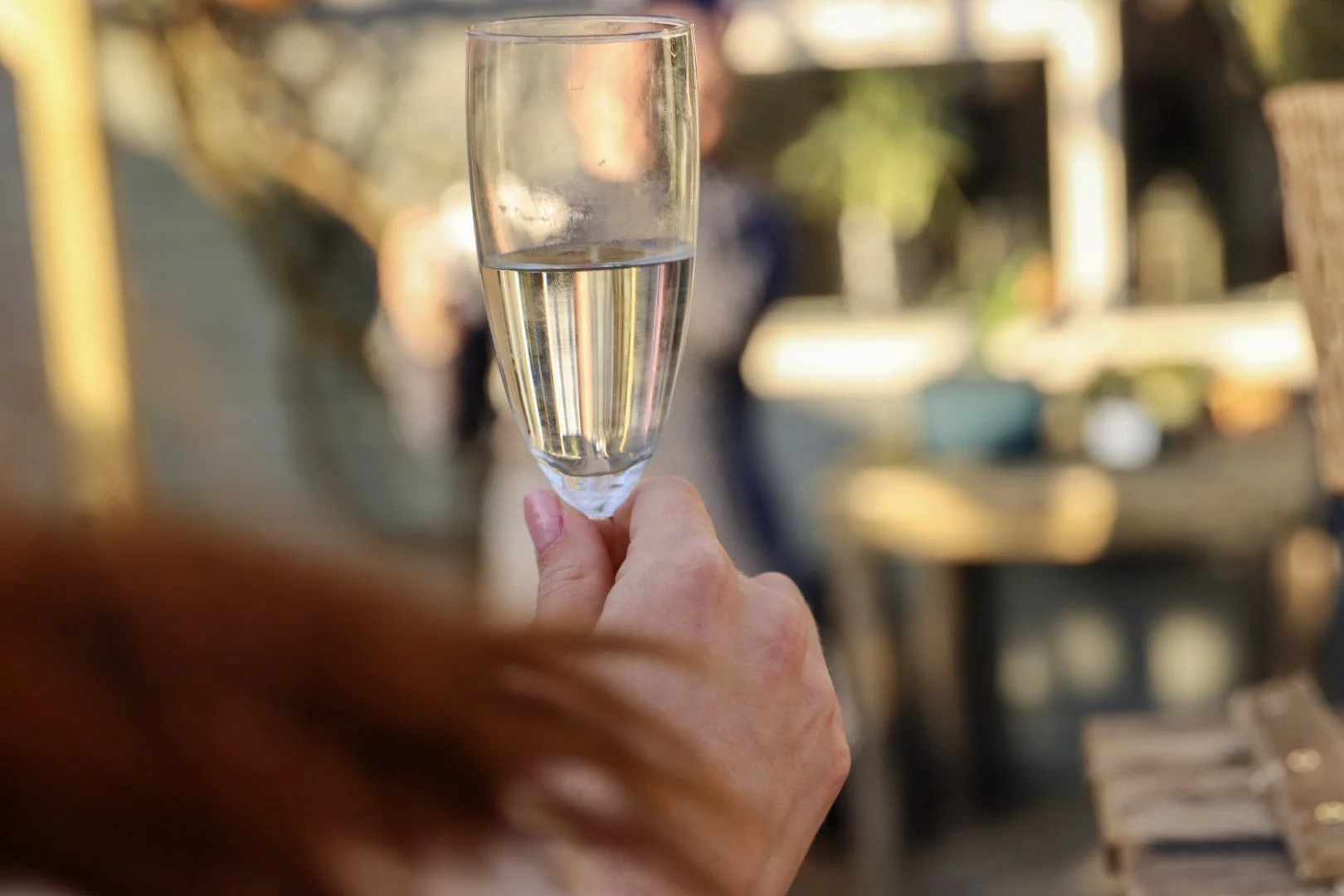 A hand holding a glass of champagne outdoors. Blurred background with people and patio furniture.