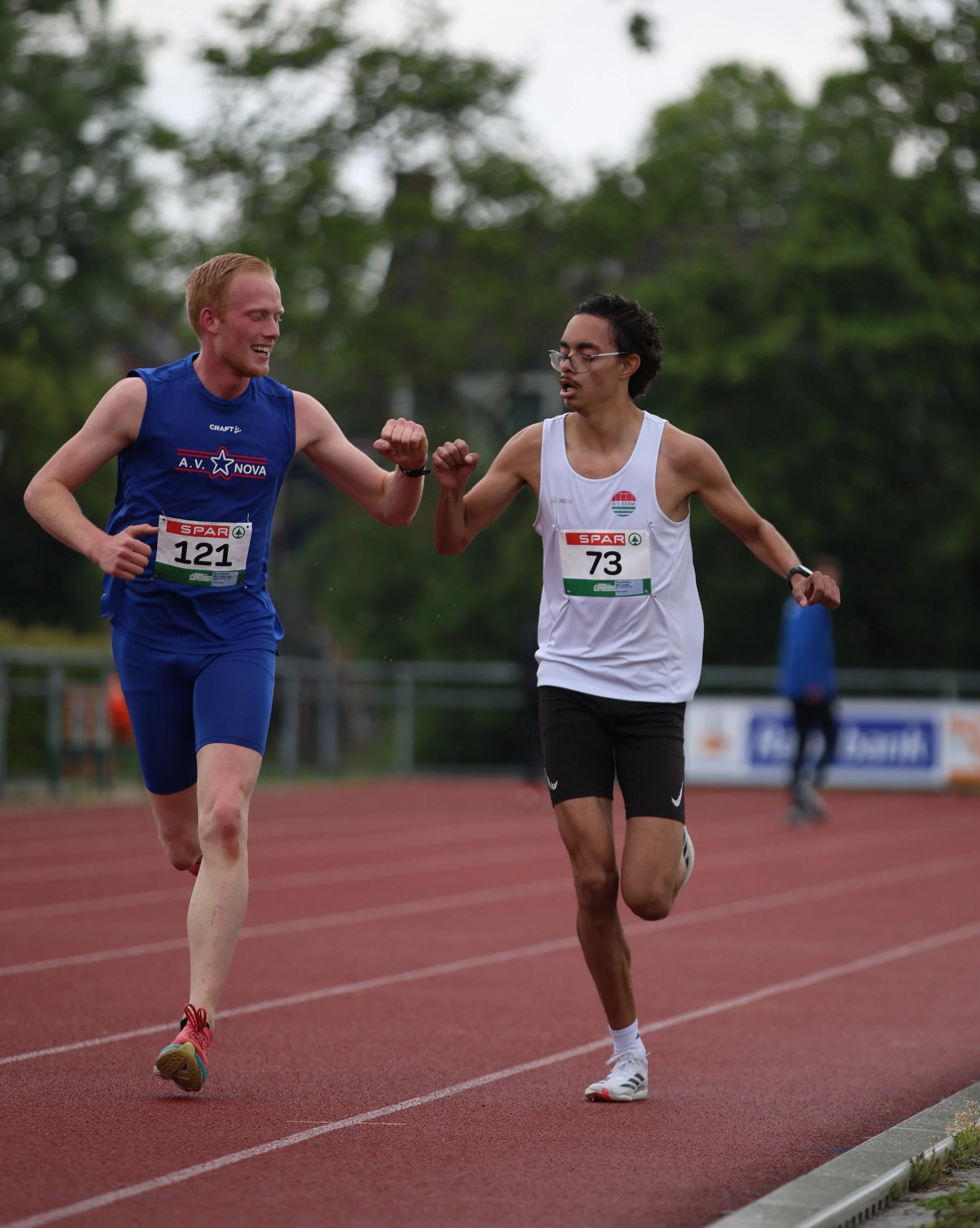 Two male runners with bib numbers 121 and 73 competing in a race on a red track, giving each other a fist bump while running outdoors with trees in the background.