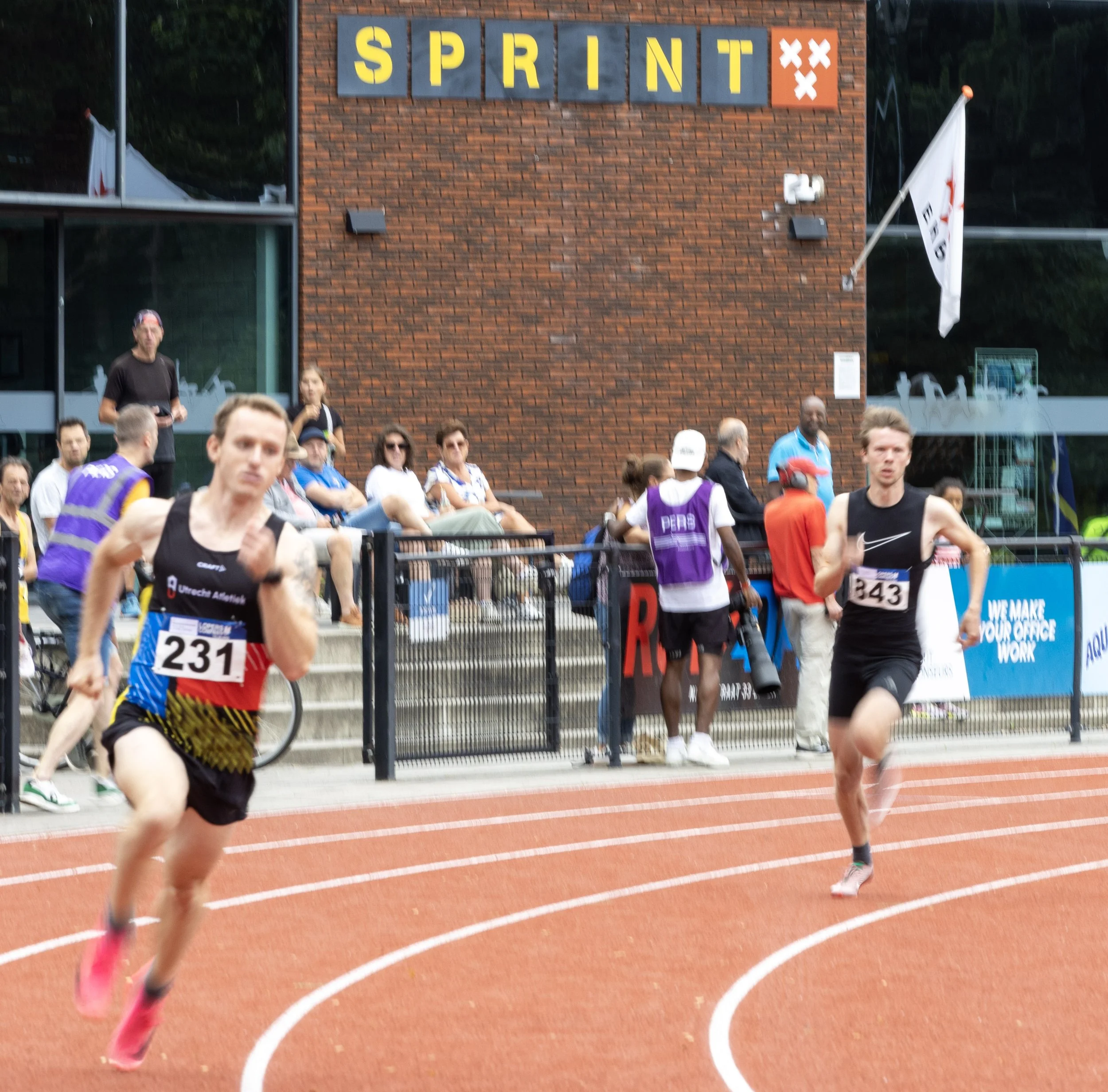 Two runners competing in a track race with spectators watching from the sidelines, behind a metal fence, outside a building with a large sign that reads 'SPRINT' on a brick wall.